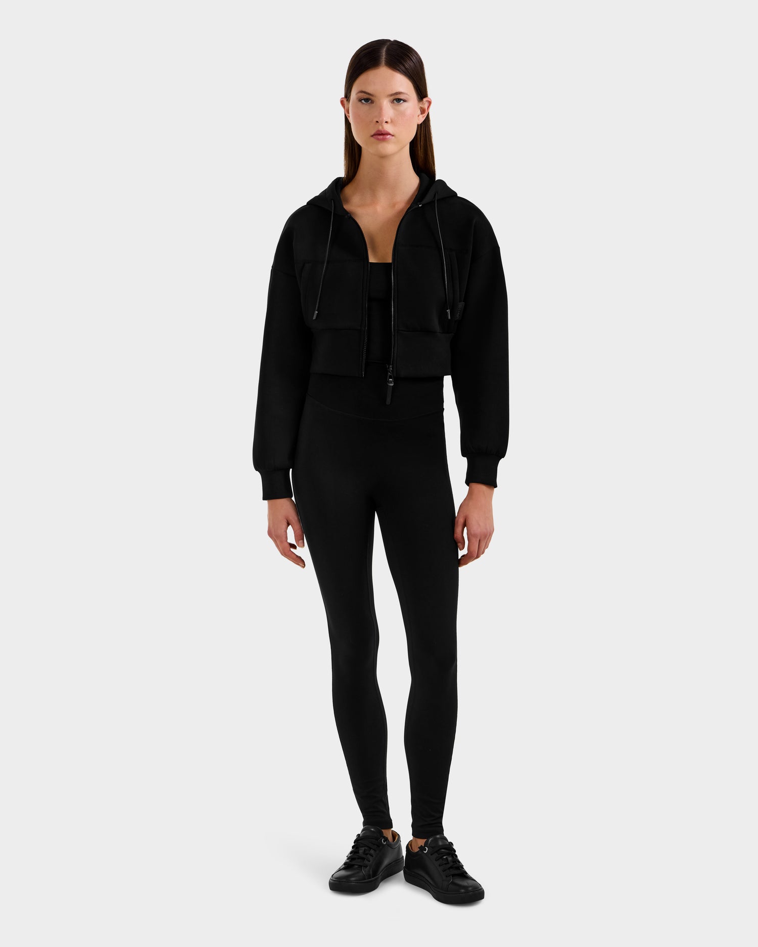 A woman faces forward against a plain white background, wearing the Rudsak ALAYA - 8525038 Black women’s hoodie over a black crop top, styled with black leggings and sneakers for an effortless casual-chic loungewear look.