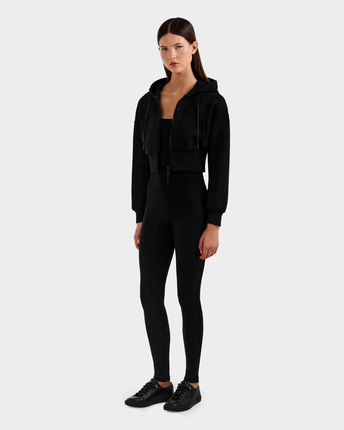 A woman poses against a light gray background wearing the Rudsak ALAYA - 8525038 Black set: an all-black cropped zip-up hoodie, fitted top, and high-waisted leggings for a casual-chic loungewear look.