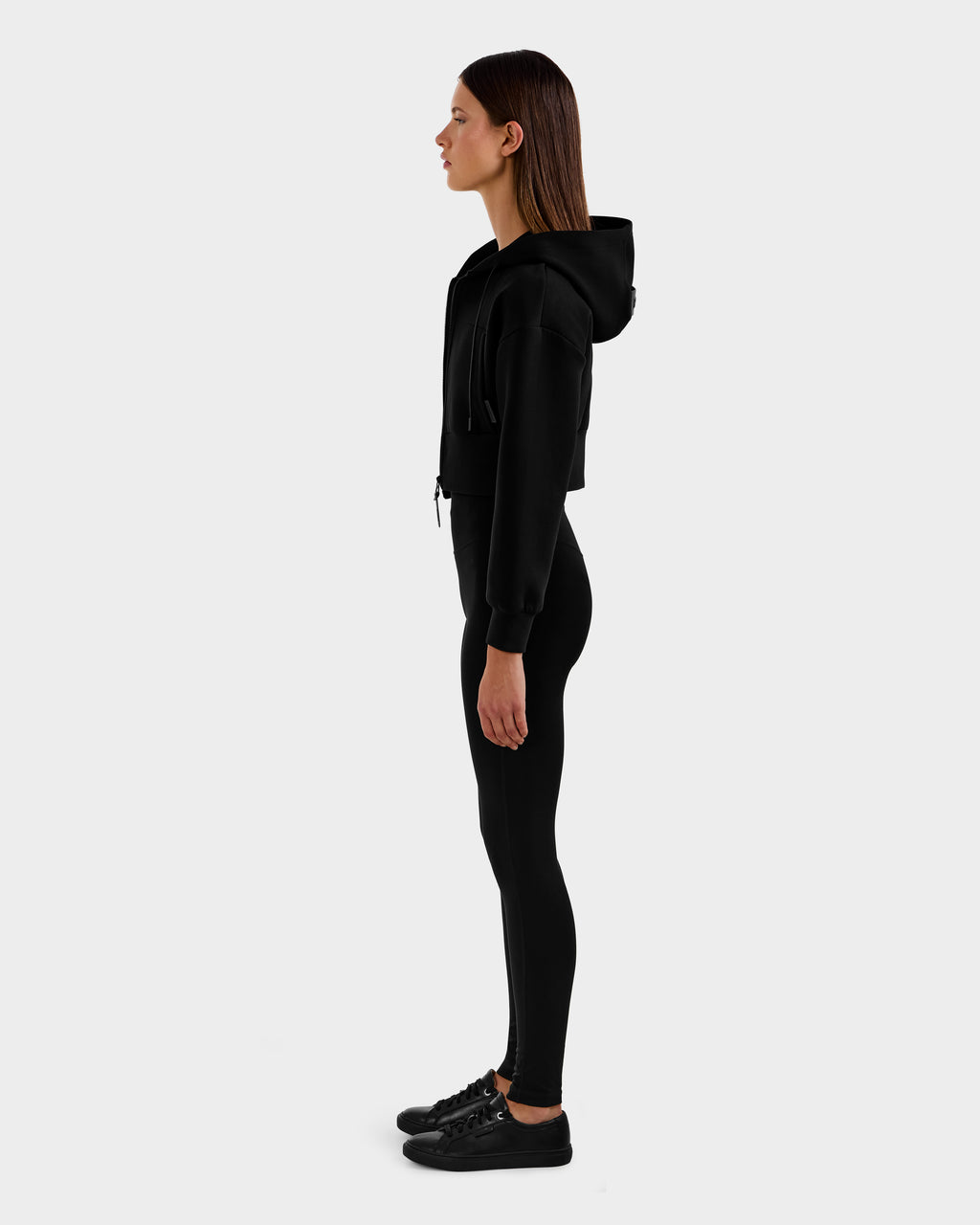 A woman stands in profile facing left, wearing the Rudsak ALAYA - 8525038 Black zip-up hoodie, black leggings, and black sneakers against a plain light gray background.