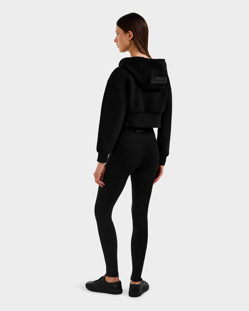 A woman models the Rudsak ALAYA - 8525038 Black cropped zip-up hoodie with black leggings and sneakers, showcasing casual-chic loungewear against a plain light gray background.