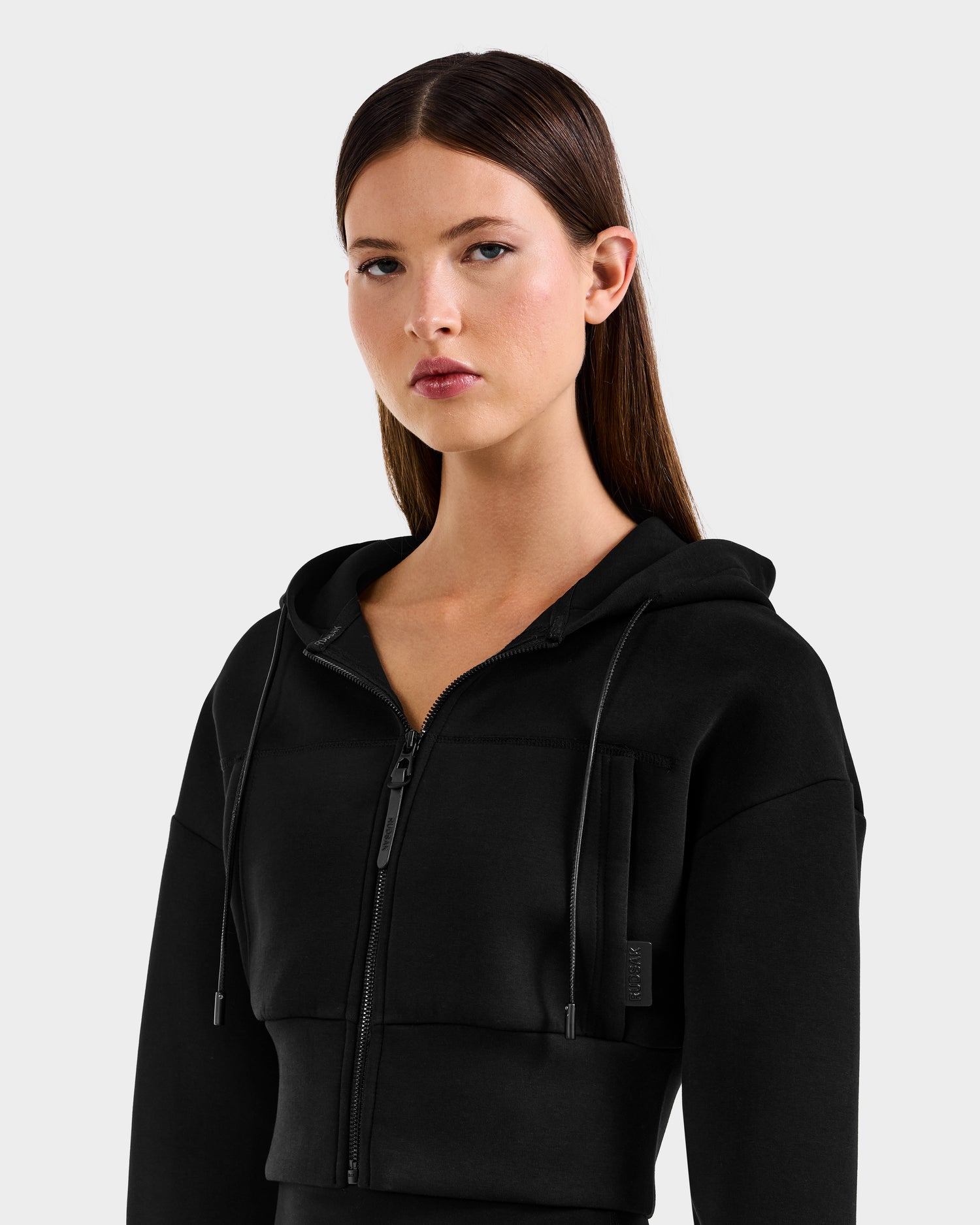 A woman with straight brown hair models the Rudsak ALAYA - 8525038 Black cropped zip-up hoodie, standing against a light gray background and looking forward with a neutral expression to highlight its casual-chic style.