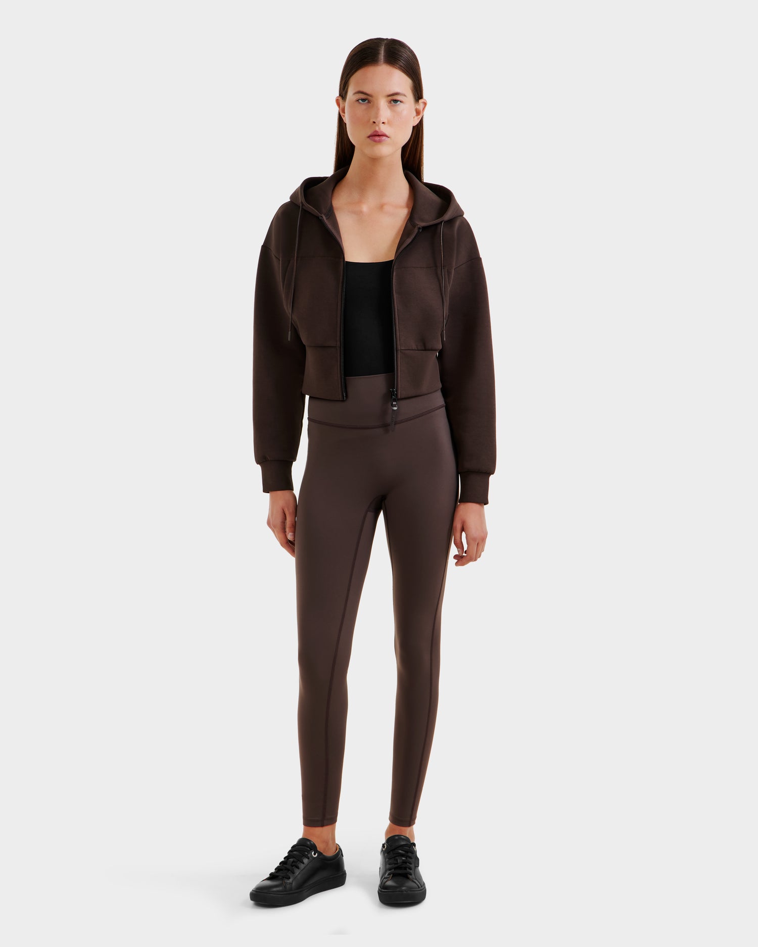 A woman faces forward in a Rudsak ALAYA - 8525038 Espresso cropped hoodie, black top, brown high-waisted leggings, and black sneakers for a casual-chic look against a plain white background.