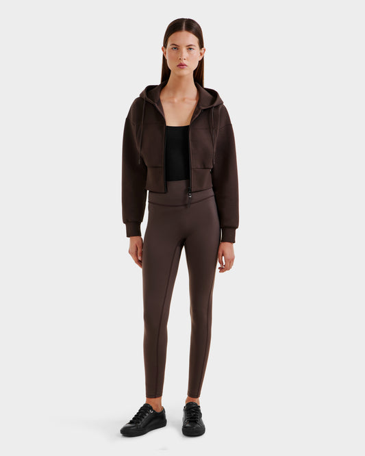 A woman faces forward in a Rudsak ALAYA - 8525038 Espresso cropped hoodie, black top, brown high-waisted leggings, and black sneakers for a casual-chic look against a plain white background.