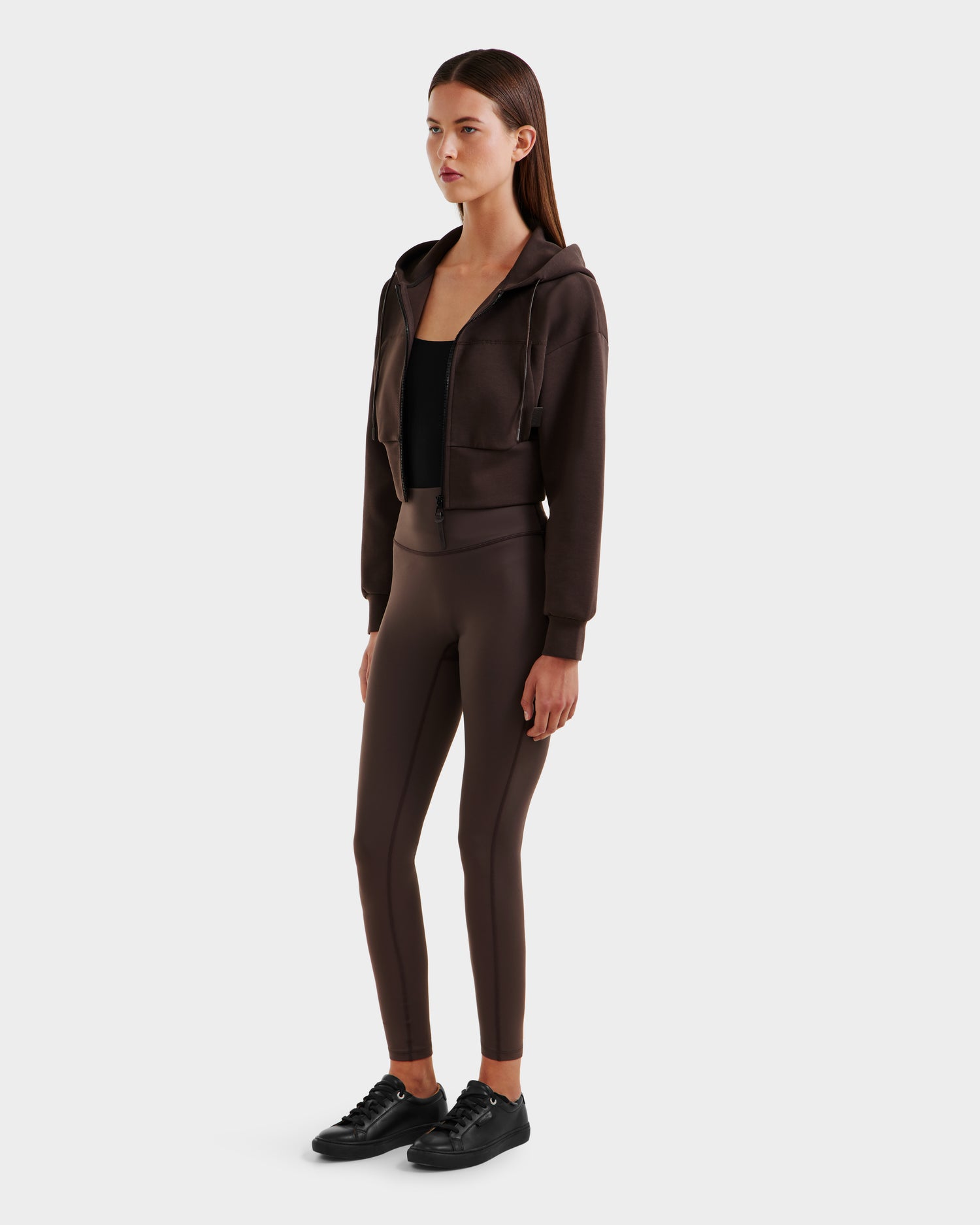 A woman with straight dark hair and a neutral expression models Rudsaks ALAYA - 8525038 Espresso hoodie against a white background, styled casually with a black top, brown leggings, and black sneakers.