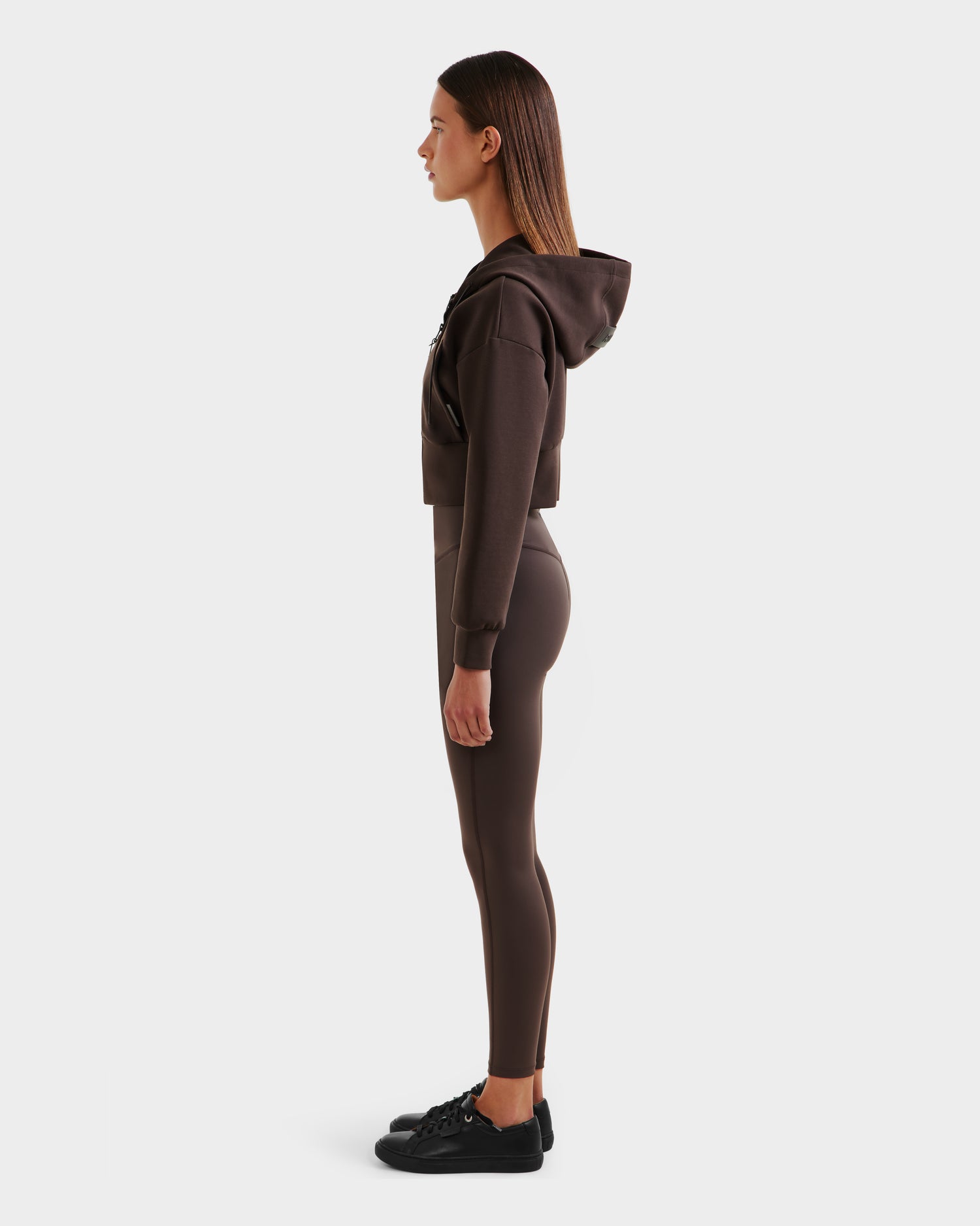 Woman in profile faces left, wearing the Rudsak ALAYA - 8525038 Espresso casual-chic brown cropped hoodie and matching leggings with black sneakers, set against a plain white background.