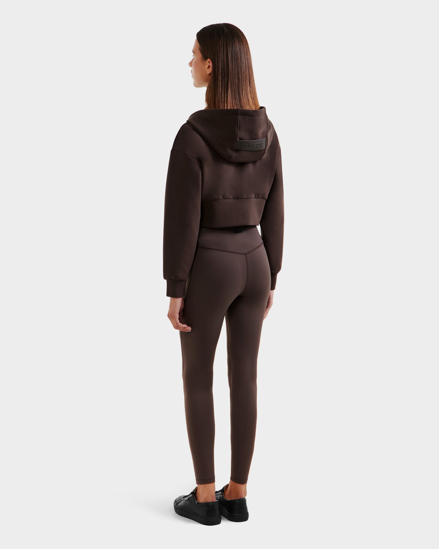 A woman faces away in Rudsak’s ALAYA - 8525038 Espresso cropped zip-up hoodie and matching leggings, paired with black sneakers. Her straight brown hair and a plain white background accentuate her casual-chic look.