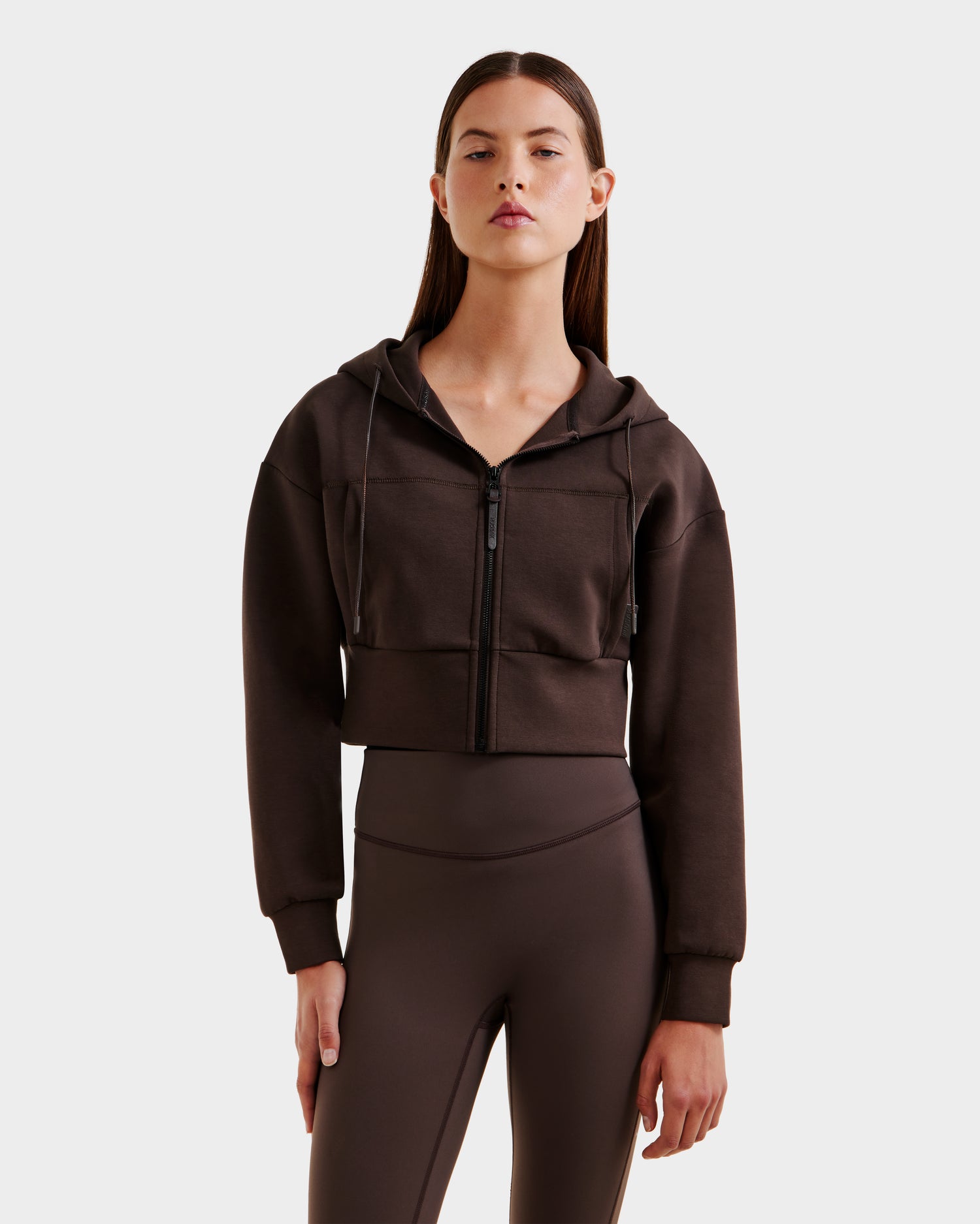 A woman with straight brown hair models the Rudsak ALAYA - 8525038 Espresso loungewear set, featuring a dark brown cropped zip-up hoodie and matching high-waisted leggings, against a plain white background.