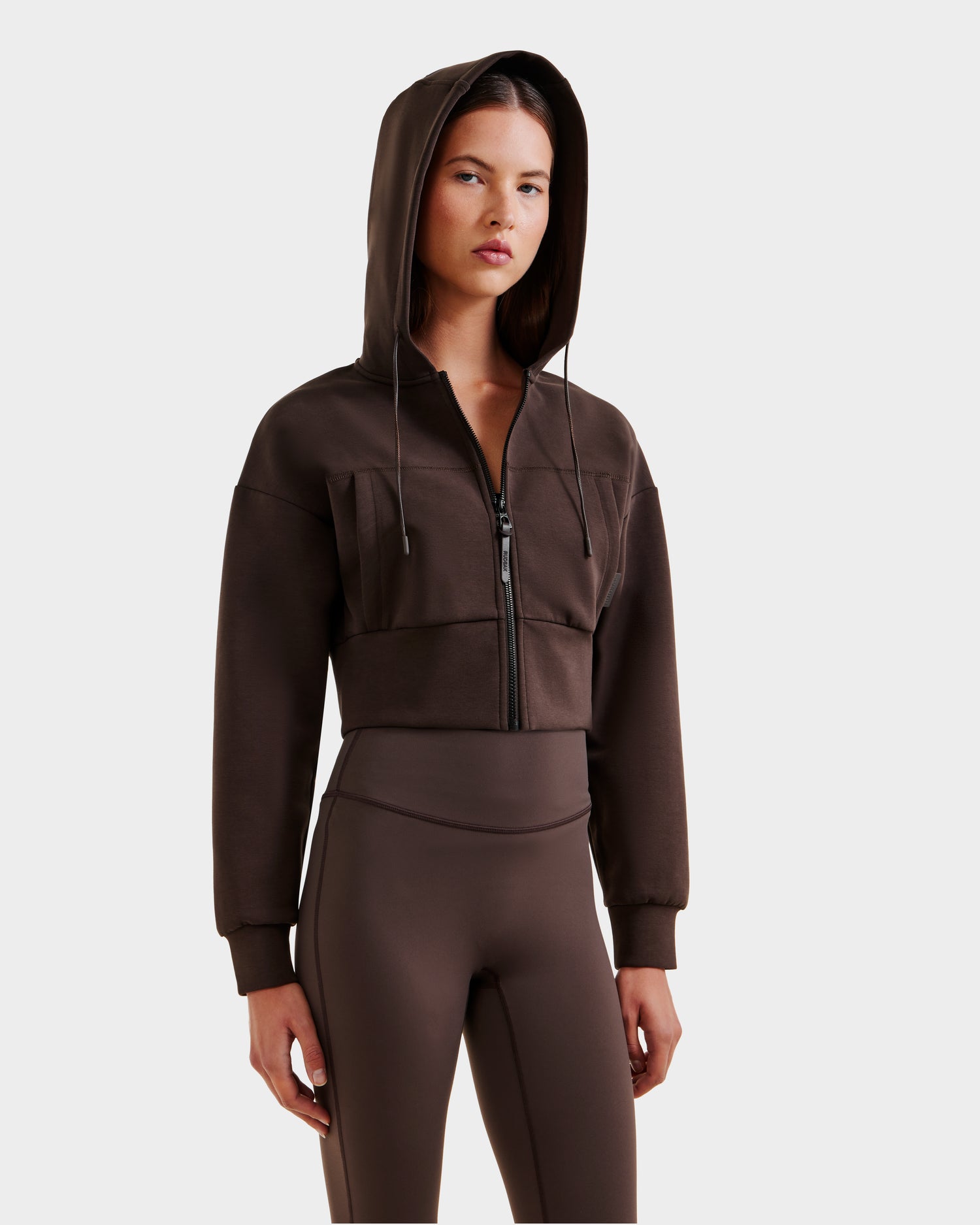 A woman wears the Rudsak ALAYA - 8525038 Espresso set— a brown cropped zip-up hoodie with matching high-waisted leggings—standing against a plain background with a neutral expression for a casual-chic look.