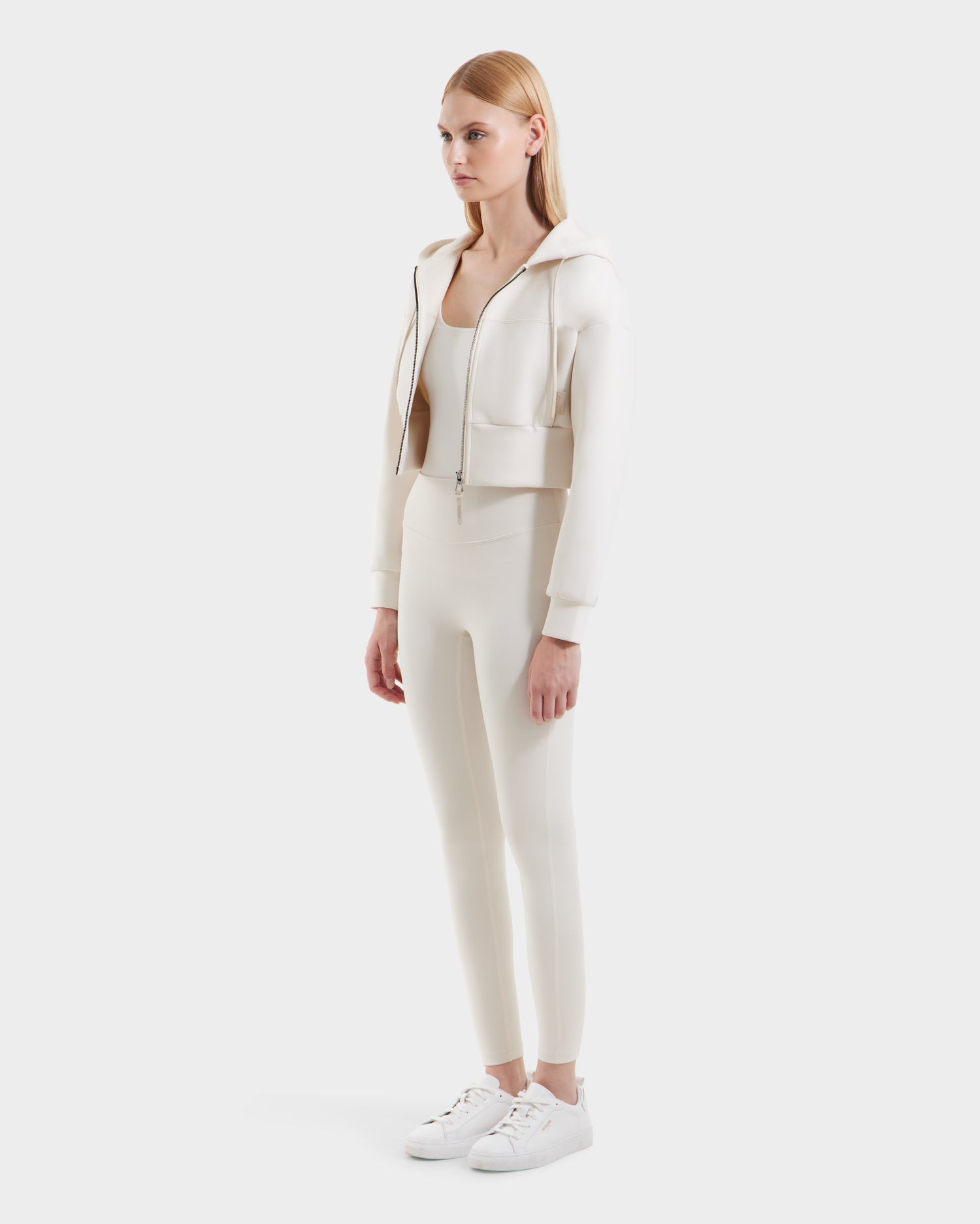 A woman faces forward in the Rudsak ALAYA - 8525038 Vanilla cropped zip-up hoodie with matching fitted leggings, tank top, and white sneakers, standing against a plain white background for a casual-chic vibe.
