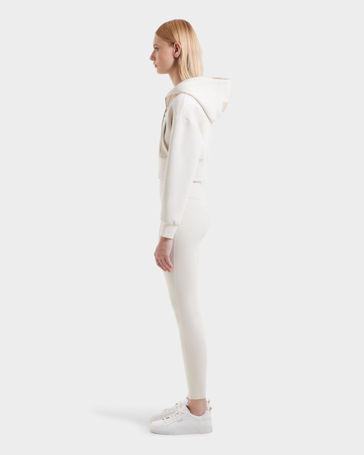 A person with straight blonde hair stands in profile wearing the Rudsak ALAYA - 8525038 Vanilla women’s cropped zip-up hoodie with matching fitted pants and white sneakers, creating a casual-chic look against a plain light background.