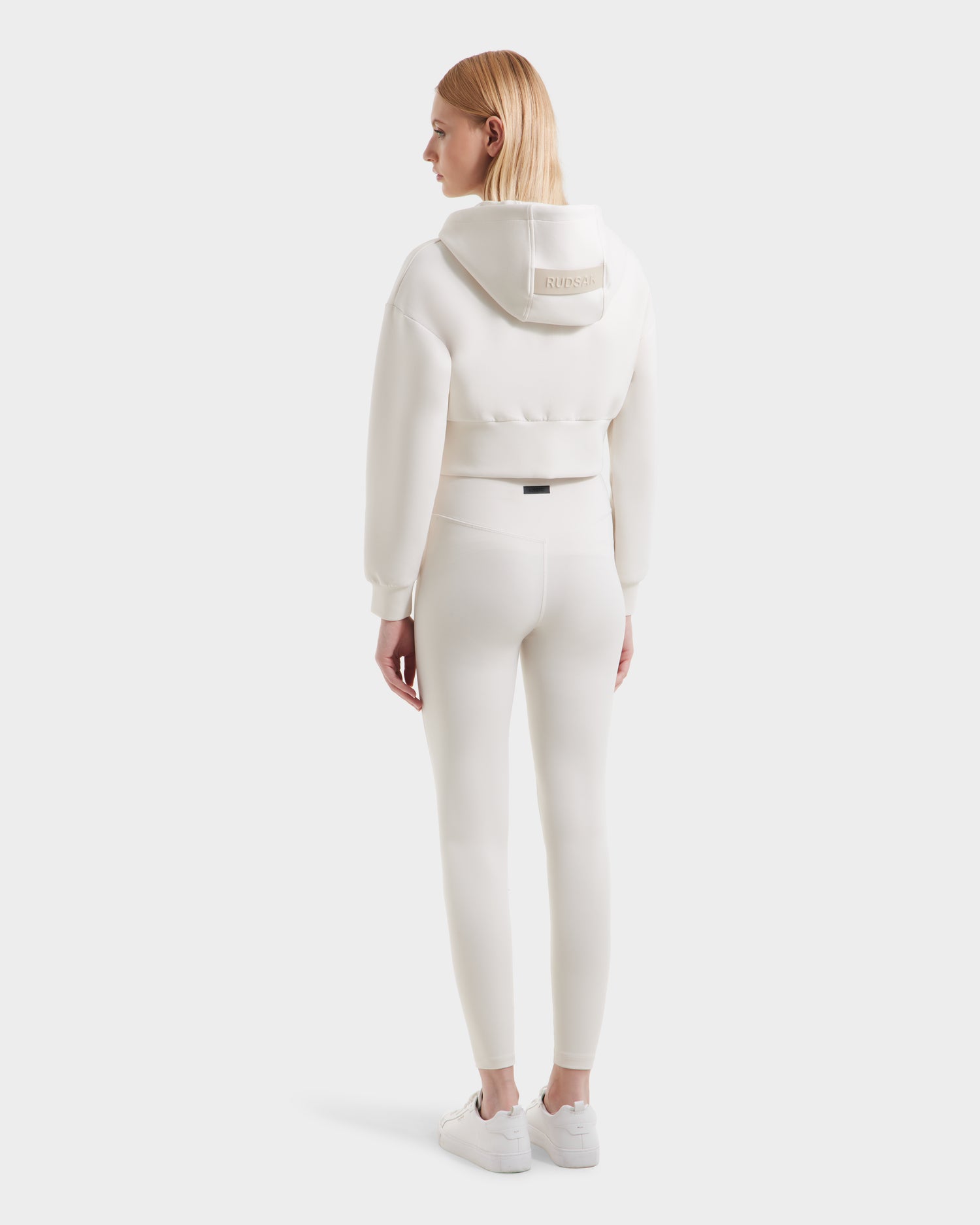 A woman with straight blonde hair and white sneakers faces away, wearing the Rudsak ALAYA - 8525038 Vanilla double jersey women’s cropped zip-up hoodie and matching high-waisted leggings against a plain light background.
