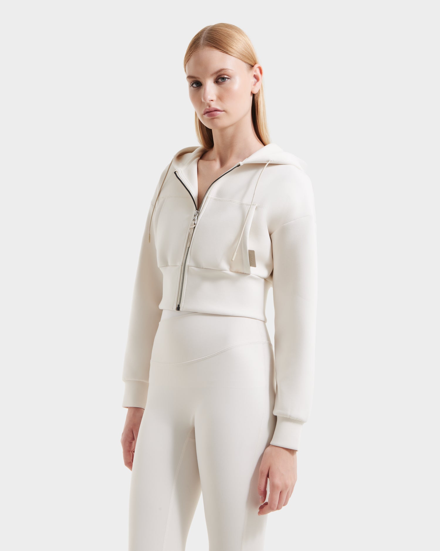 A woman with straight blonde hair wears the Rudsak ALAYA - 8525038 Vanilla cropped zip-up hoodie and matching high-waisted leggings in premium double jersey, standing against a plain light background for a casual-chic look.