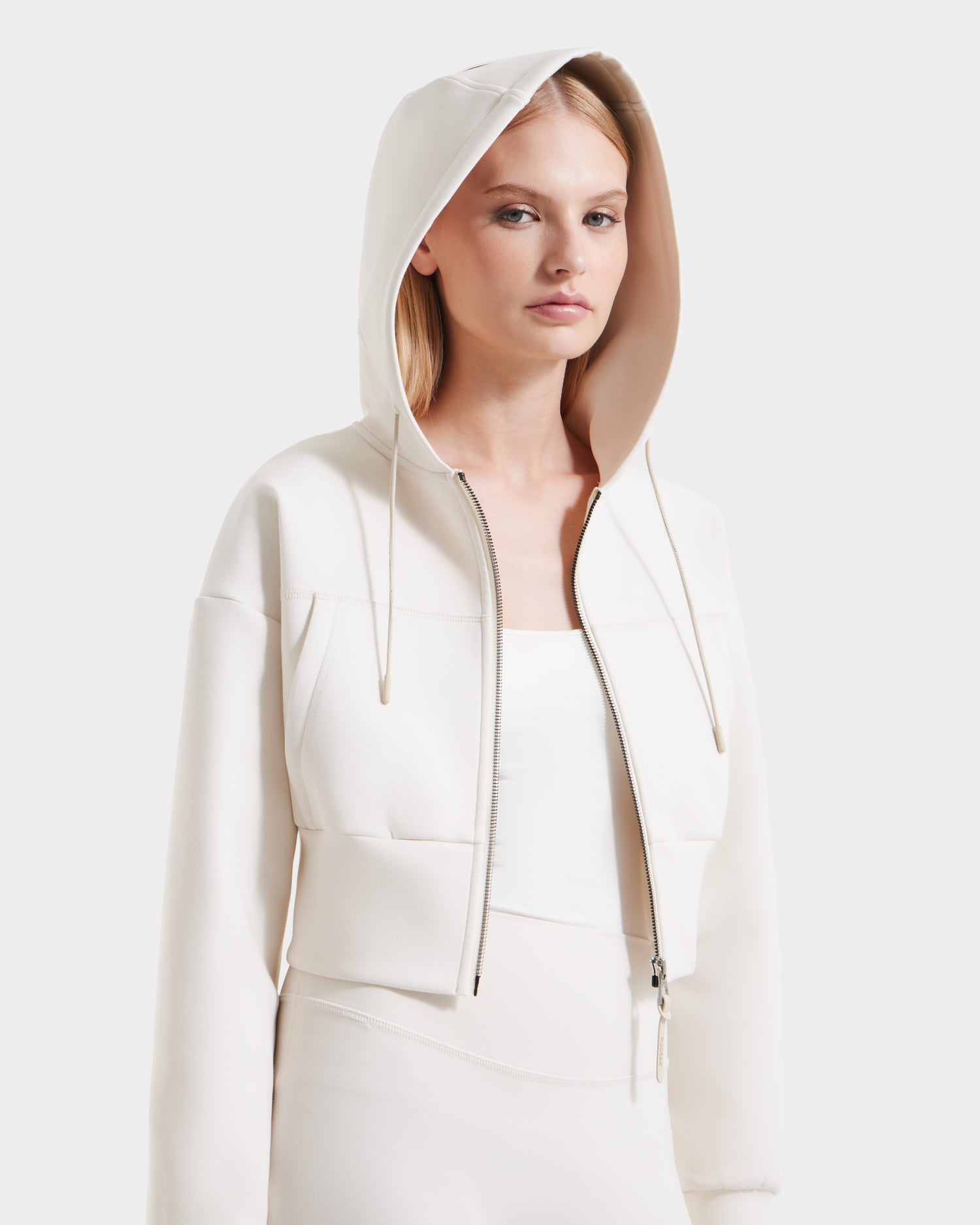 A woman with straight blonde hair models the Rudsak ALAYA - 8525038 Vanilla cropped zip-up hoodie, tank top, and skirt set in premium double jersey for a casual-chic look against a plain white background.