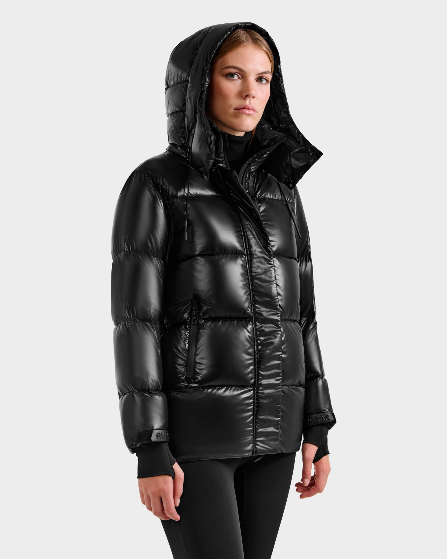 A woman wearing the Rudsak ALESSANDRA S-S - L125560 Black down puffer with a hood stands against a light gray background, gazing slightly to the side with a neutral expression.