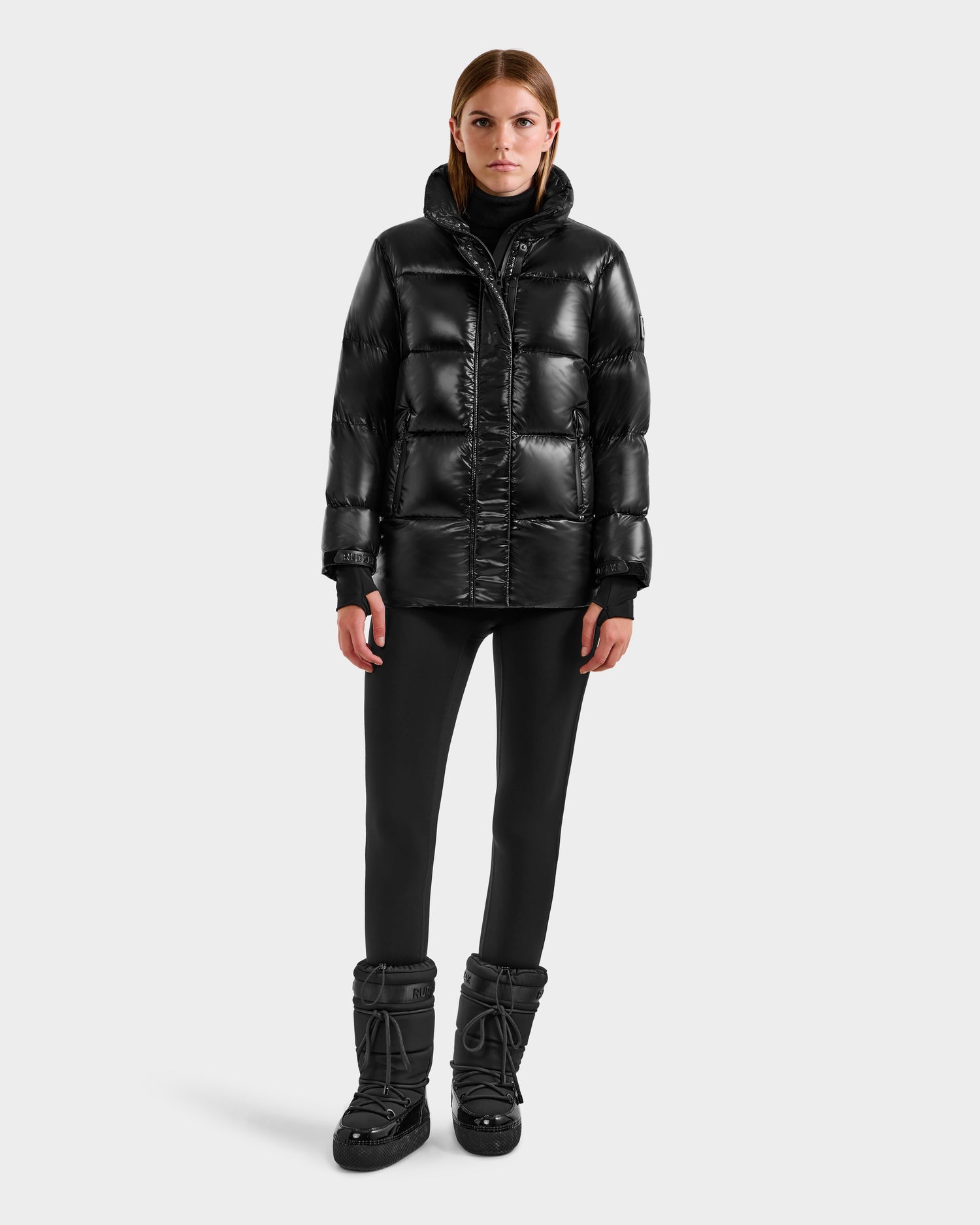 A woman faces forward against a light gray background, wearing the Rudsak ALESSANDRA S-S - L125560 Black glossy women’s down puffer jacket, paired with black pants and winter boots.
