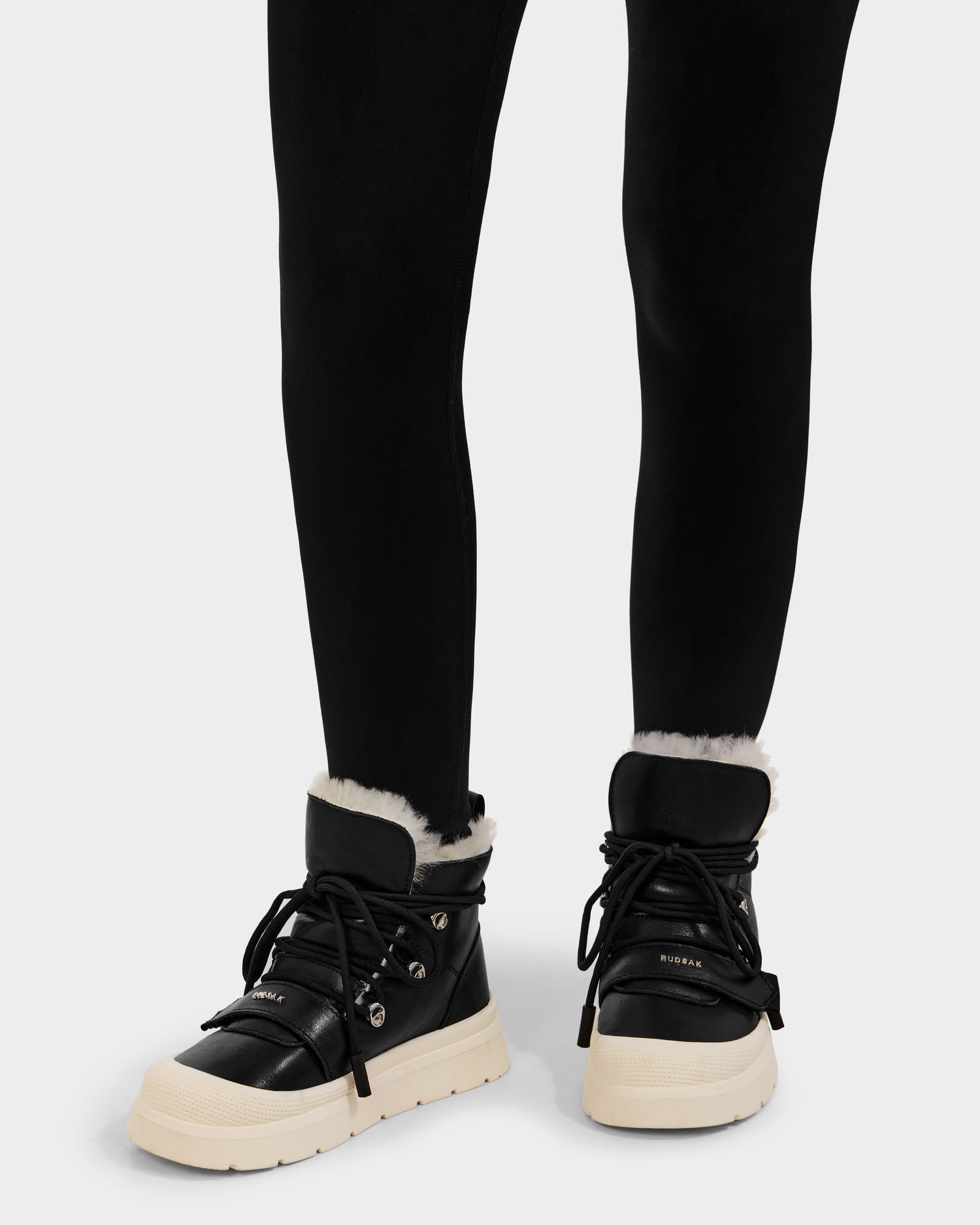 A person wearing Rudsak ALEXEY L - L225518 Black winter boots with white soles and visible wool lining, paired with black leggings, standing against a plain background.