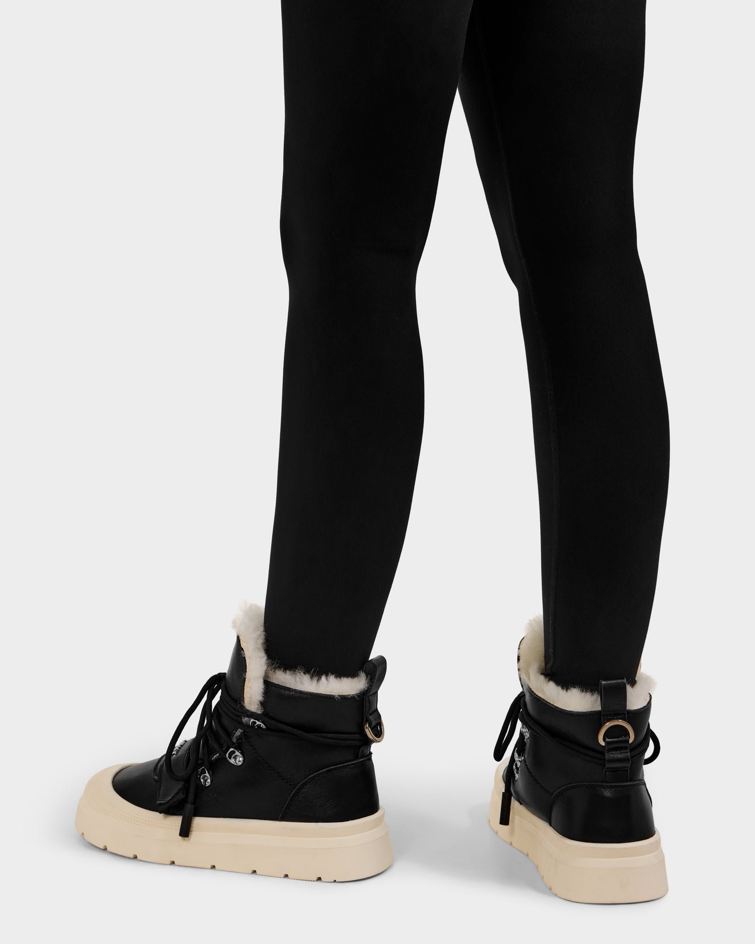 A person wearing black leggings and Rudsak’s ALEXEY L - L225518 Black women’s winter boots with white wool lining and beige chunky soles, shown from the back against a plain light background.