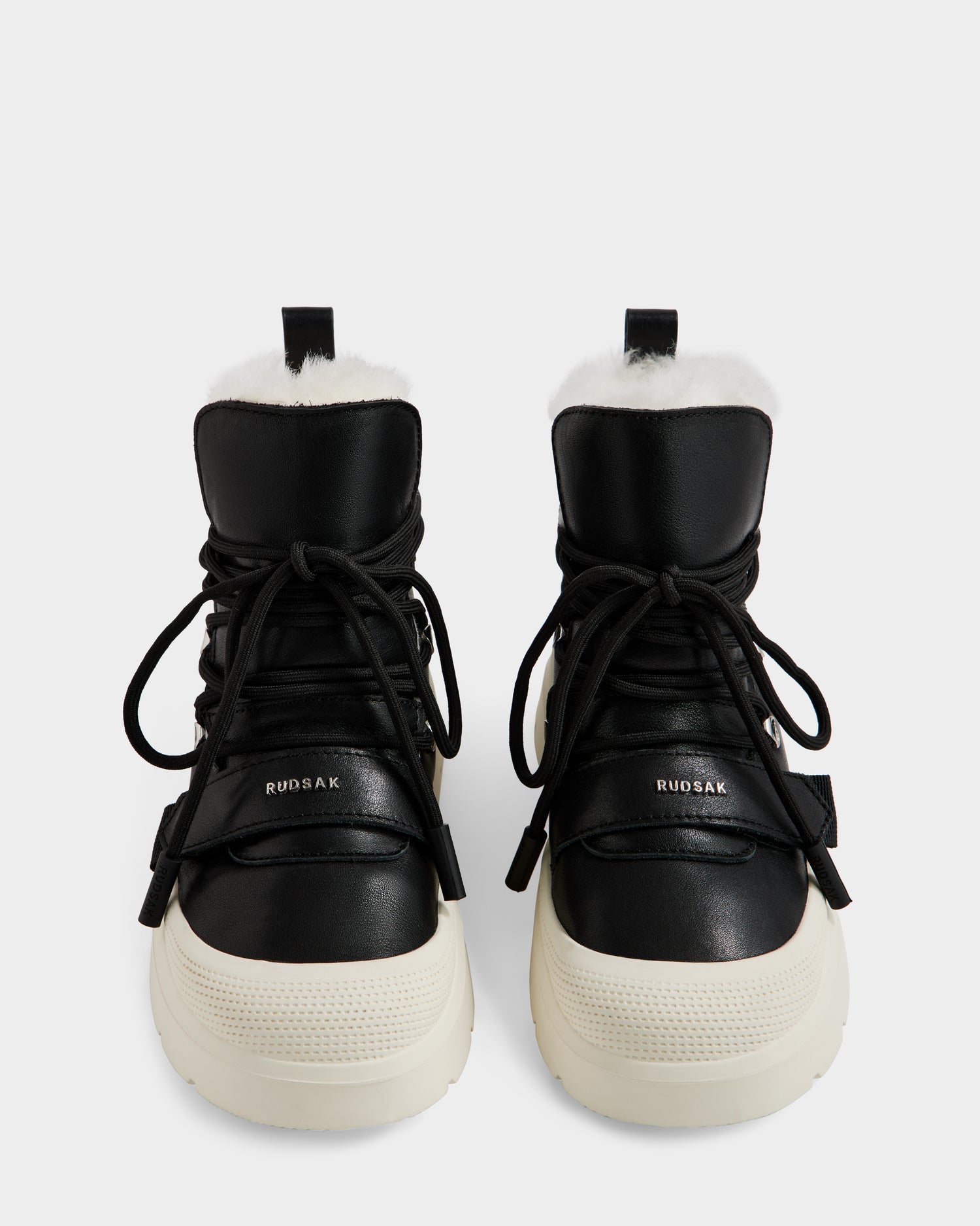 A pair of Rudsak ALEXEY L - L225518 Black women’s leather winter boots with thick white soles, black laces, and visible plush wool lining, shown from the front on a plain white background.