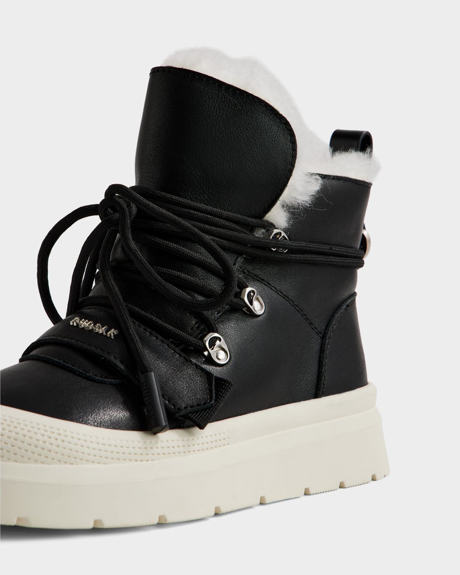 A close-up of the Rudsak ALEXEY L - L225518 Black women’s leather winter boot featuring a thick white sole, silver eyelets, black laces, and plush white faux fur lining visible at the top.