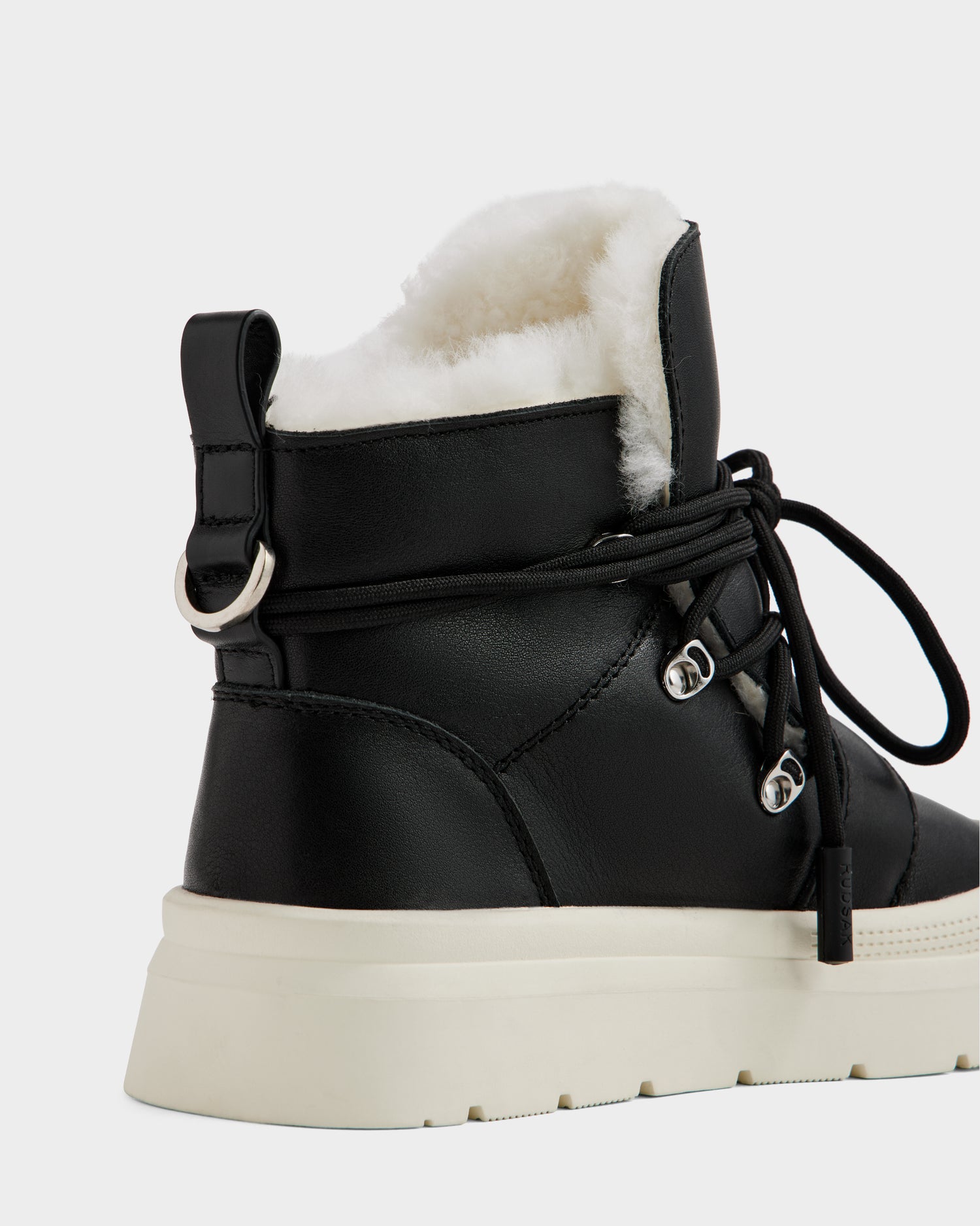 A close-up of the Rudsak ALEXEY L - L225518 Black women’s leather boot featuring black lace-up detail, plush white wool lining, and a thick white sole, shown from the back and side against a plain background.