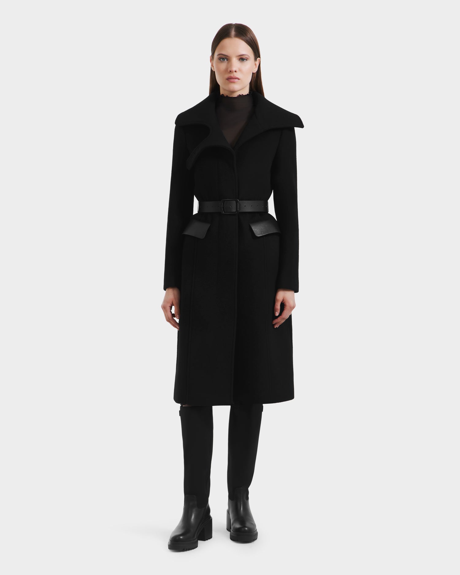 A woman faces forward in a long, fitted ALICE - 8125606 Black wool coat by Rudsak with a high collar, black pants, and black ankle boots, standing against a plain white background.