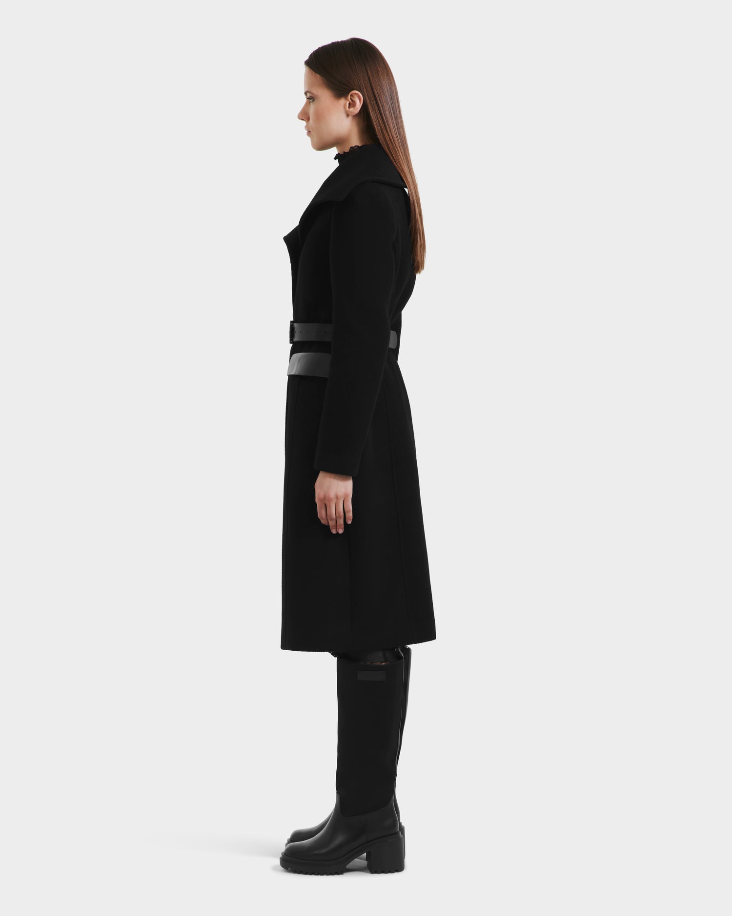 A woman in profile stands against a white background, wearing the Rudsak ALICE - 8125606 Black womens wool coat with a belt, black pants, and knee-high black boots. Her straight brown hair falls past her shoulders.