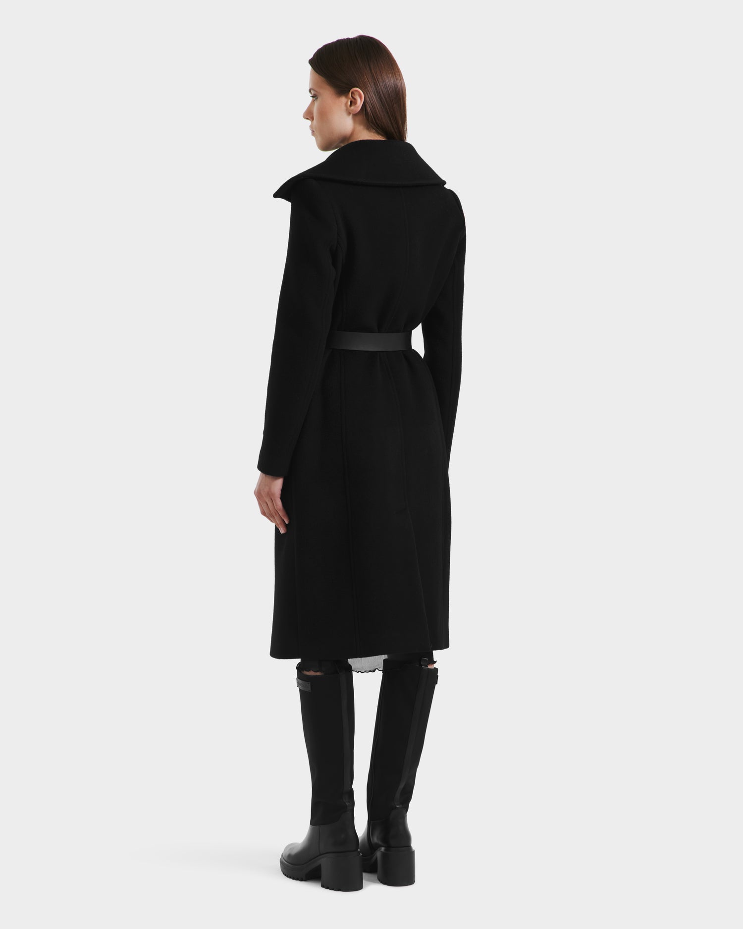 A woman with straight brown hair wears the Rudsak ALICE - 8125606 Black fitted wool coat and black knee-high boots, standing against a plain white background and looking to her left.