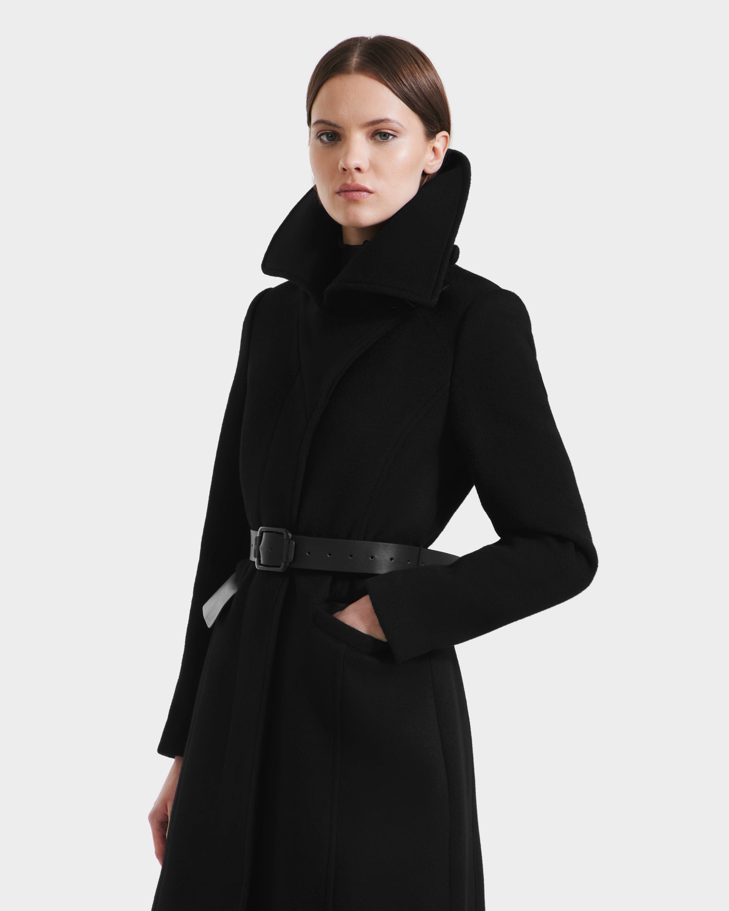 A woman with straight brown hair models the Rudsak ALICE - 8125606 Black, a high-collared, belted fitted wool coat, standing with one hand in her pocket against a plain white background.