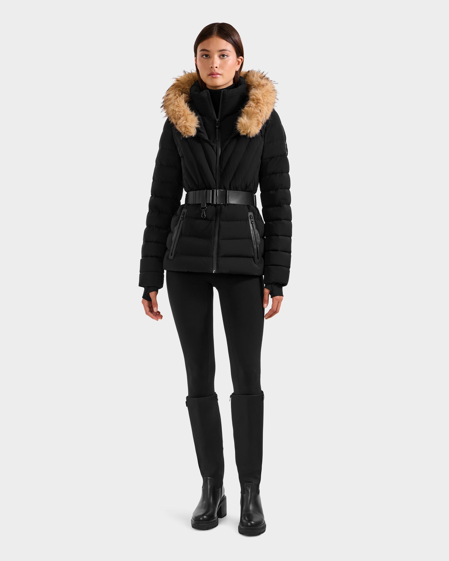 A woman faces forward wearing the Rudsak ALISA F - L125527 Black quilted puffer jacket with a faux-fur trim hood, paired with black leggings and boots, set against a plain light gray background.