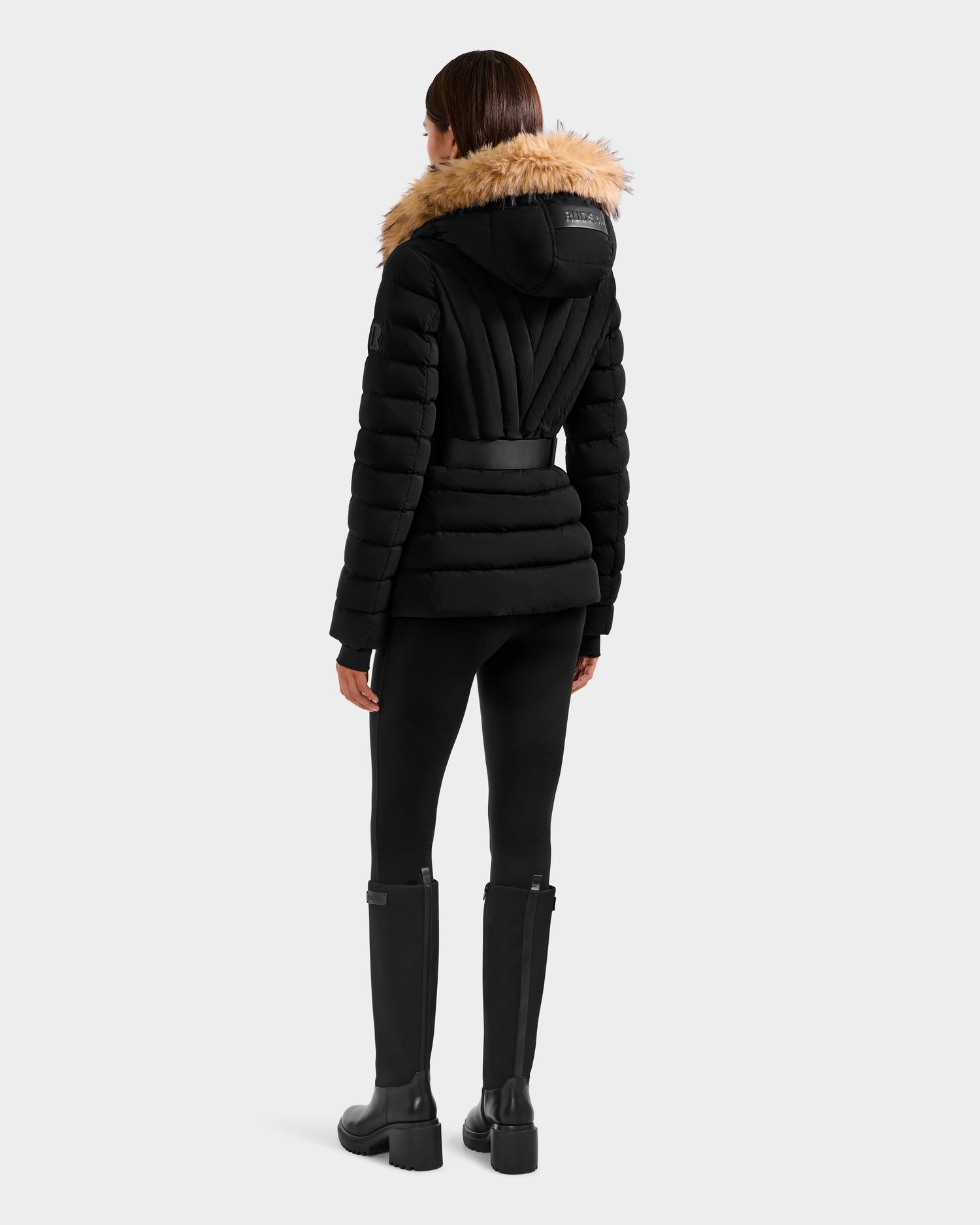 A woman faces away, wearing the Rudsak ALISA F - L125527 Black quilted puffer jacket with a faux-fur trim hood, paired with black leggings and tall black boots. The background is plain light gray.
