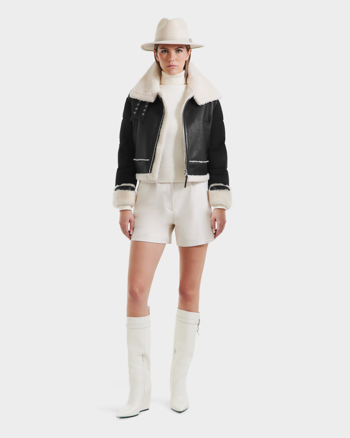 A woman faces forward against a white background, wearing the Rudsak ANA - 8125508 Black women’s shearling jacket with leather trim, cream hat, turtleneck sweater, cream shorts, and knee-high cream boots.