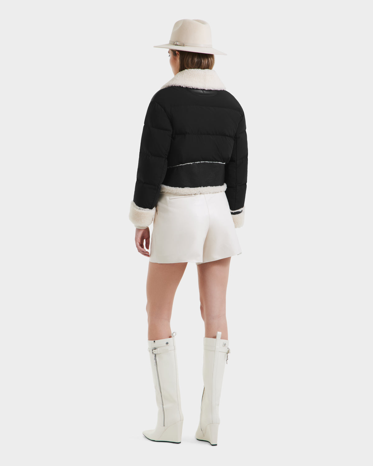 A person faces away, wearing the Rudsak ANA - 8125508 Black cropped puffer jacket with leather trim, paired with a white mini skirt, tall zippered white boots, and a wide-brimmed beige hat against a plain background.