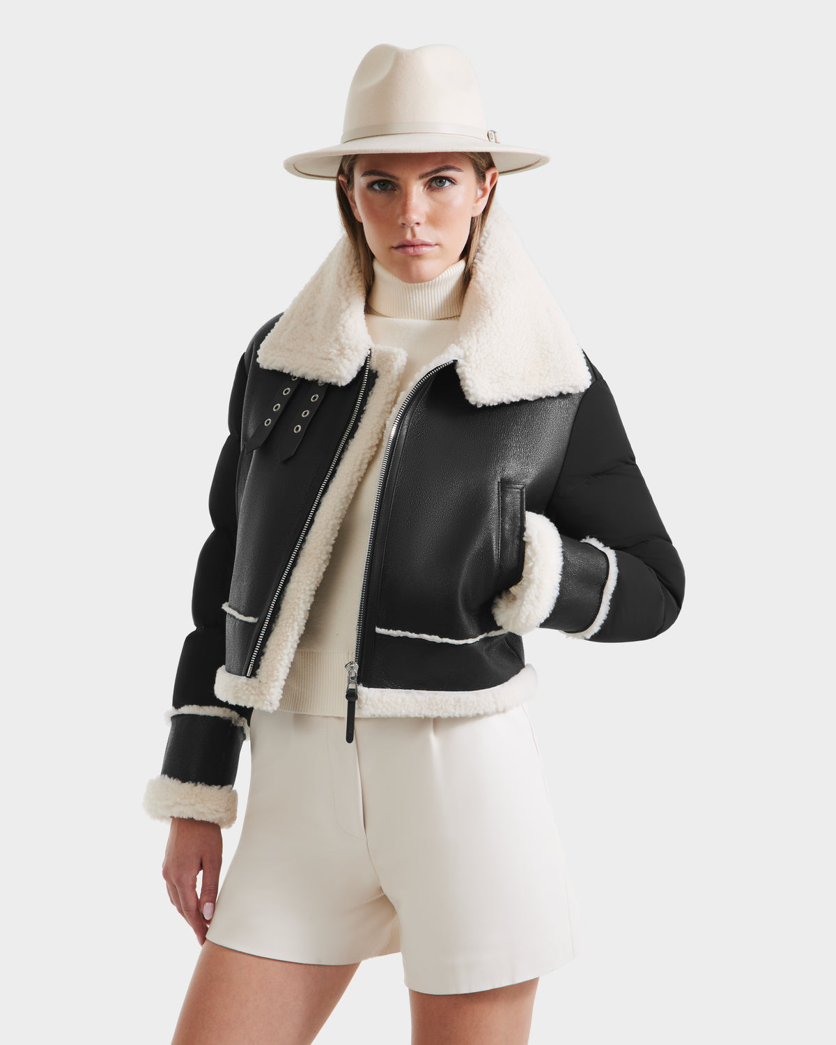 A woman models the Rudsak ANA - 8125508 Black shearling jacket, featuring white fur trim, paired with a cream wide-brim hat, turtleneck sweater, and high-waisted shorts against a plain white background.