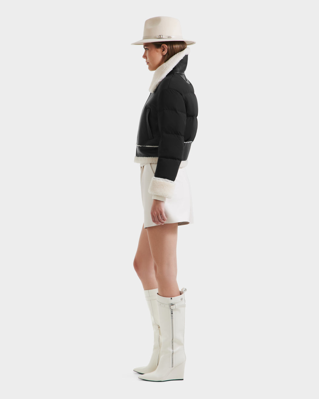 A woman in profile wears the Rudsak ANA - 8125508 Black women’s shearling jacket with leather trim, paired with a white wide-brim hat, white shorts, and knee-high white wedge boots against a plain light background.