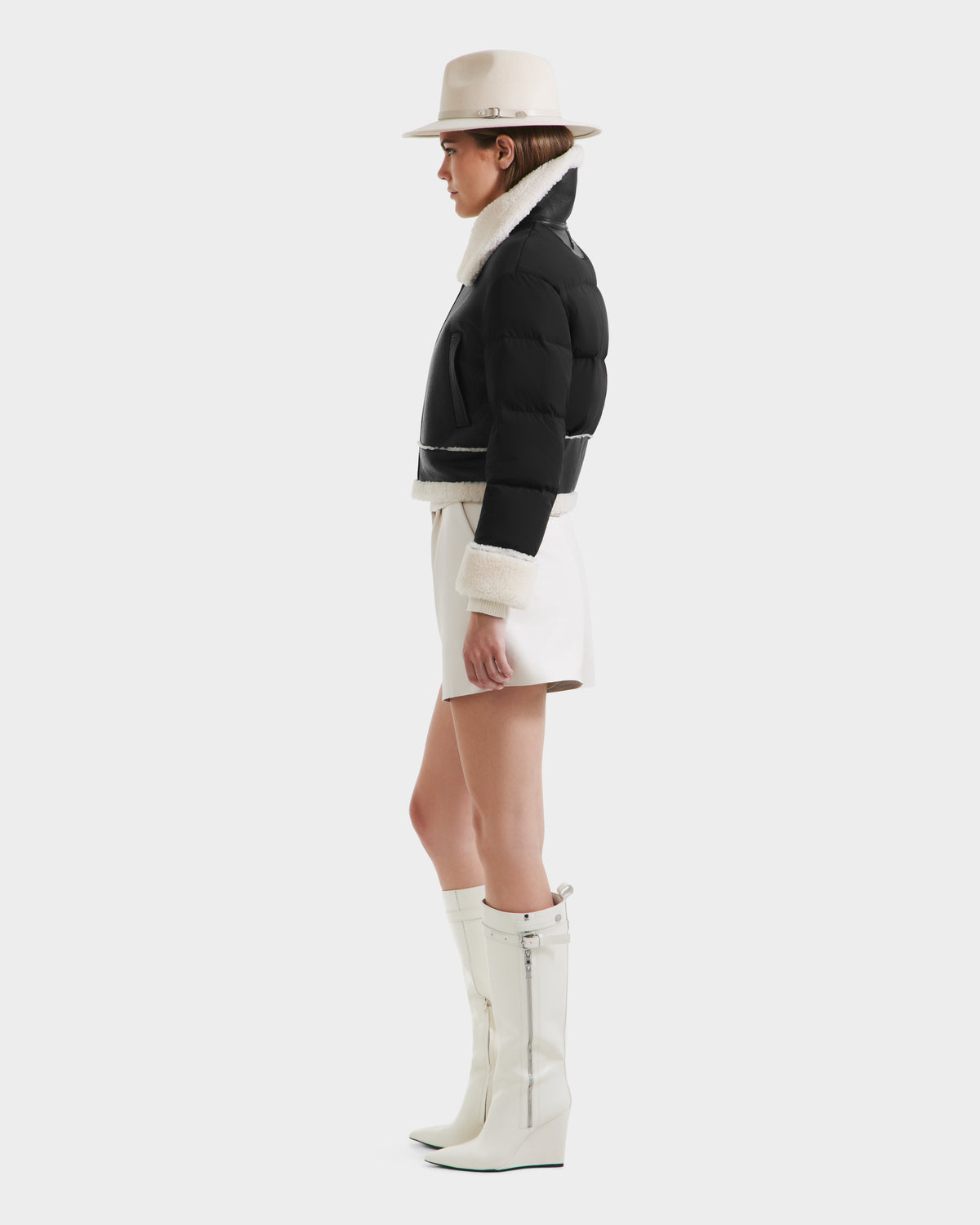 A woman in profile wears the Rudsak ANA - 8125508 Black women’s shearling jacket with leather trim, paired with a white wide-brim hat, white shorts, and knee-high white wedge boots against a plain light background.