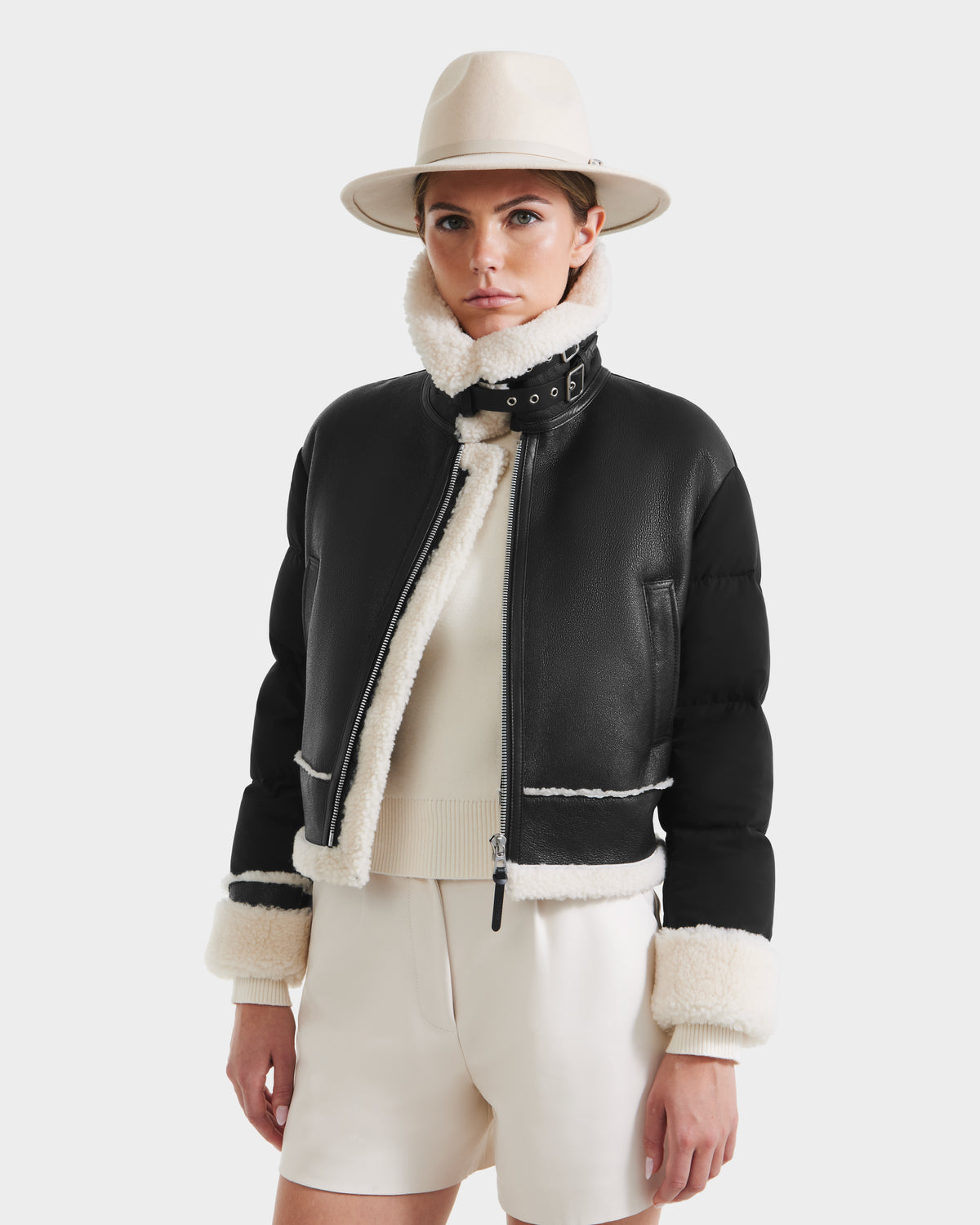 A woman in the Rudsak ANA - 8125508 Black women’s shearling jacket with white fleece trim, a cream sweater, and light shorts stands against a plain white background, wearing a light-colored hat.