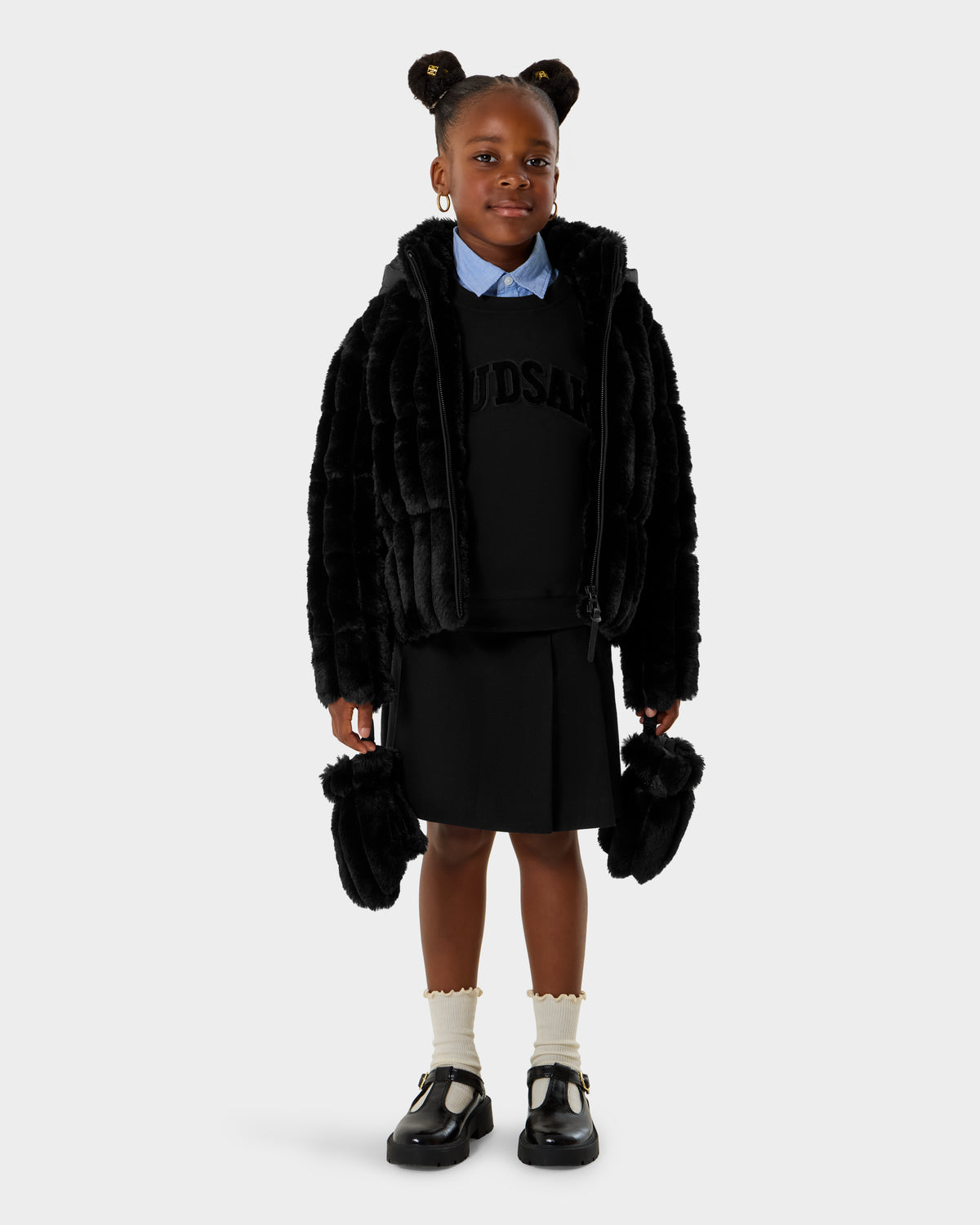 A young girl smiles forward, wearing the Rudsak ANASTASIA K - 0125000 Black faux fur shell jacket, with a black skirt, text sweater, blue collared shirt, white socks, black shoes, and holding a small black fur bag. Her hair is styled in two buns.