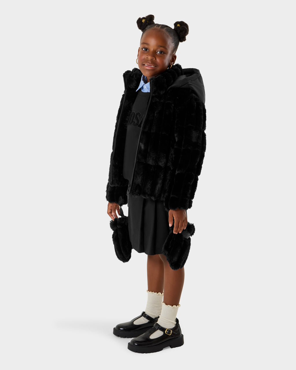 A young girl wearing the Rudsak ANASTASIA K - 0125000 Black faux fur shell jacket stands smiling at the camera. She pairs it with a black dress, cream socks, black Mary Jane shoes, and her hair in two buns against a plain light background.