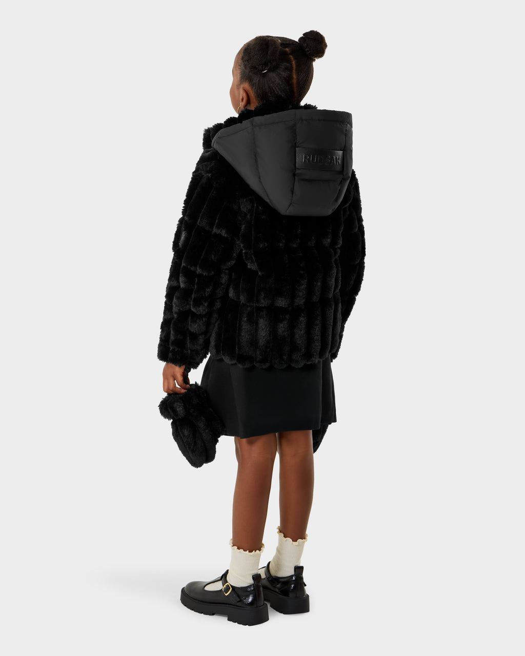 A young girl with her hair in a bun wears the Rudsak ANASTASIA K - 0125000 Black puffer with a faux fur shell and hood, black skirt, ruffled white socks, black shoes, and a matching furry bag against a plain background.