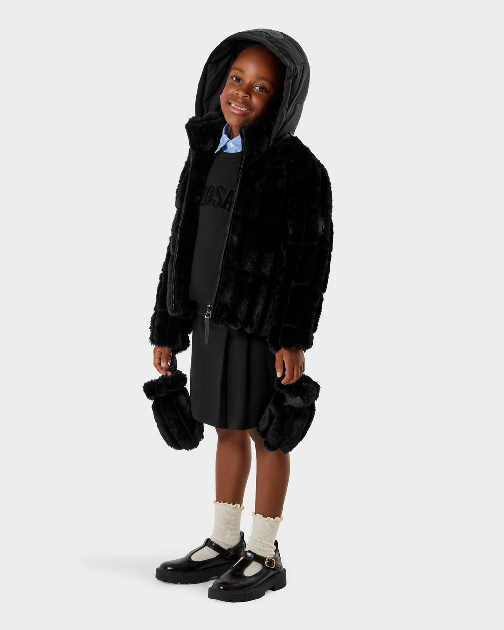 A young child smiles in the Rudsak ANASTASIA K - 0125000 Black faux fur shell hooded jacket with matching mittens, black dress, white socks, and black strap shoes. The background is plain white.