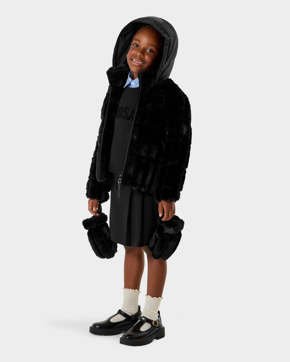 A young child smiles in the Rudsak ANASTASIA K - 0125000 Black faux fur shell hooded jacket with matching mittens, black dress, white socks, and black strap shoes. The background is plain white.
