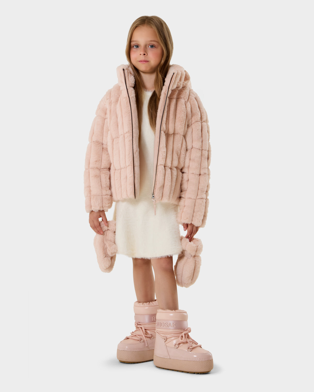 A young girl with long hair wears the Rudsak ANASTASIA K - 0125000 Pastel Pink puffer jacket, a white dress, and oversized pink boots. She holds matching mittens and stands against a plain light background, facing the camera.