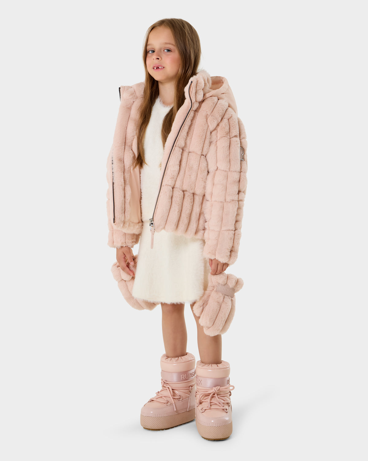 A young girl faces forward against a plain background, wearing the Rudsak ANASTASIA K - 0125000 Pastel Pink hooded fur jacket with a white dress and matching pink snow boots, holding a small pink purse. She has long, straight hair.