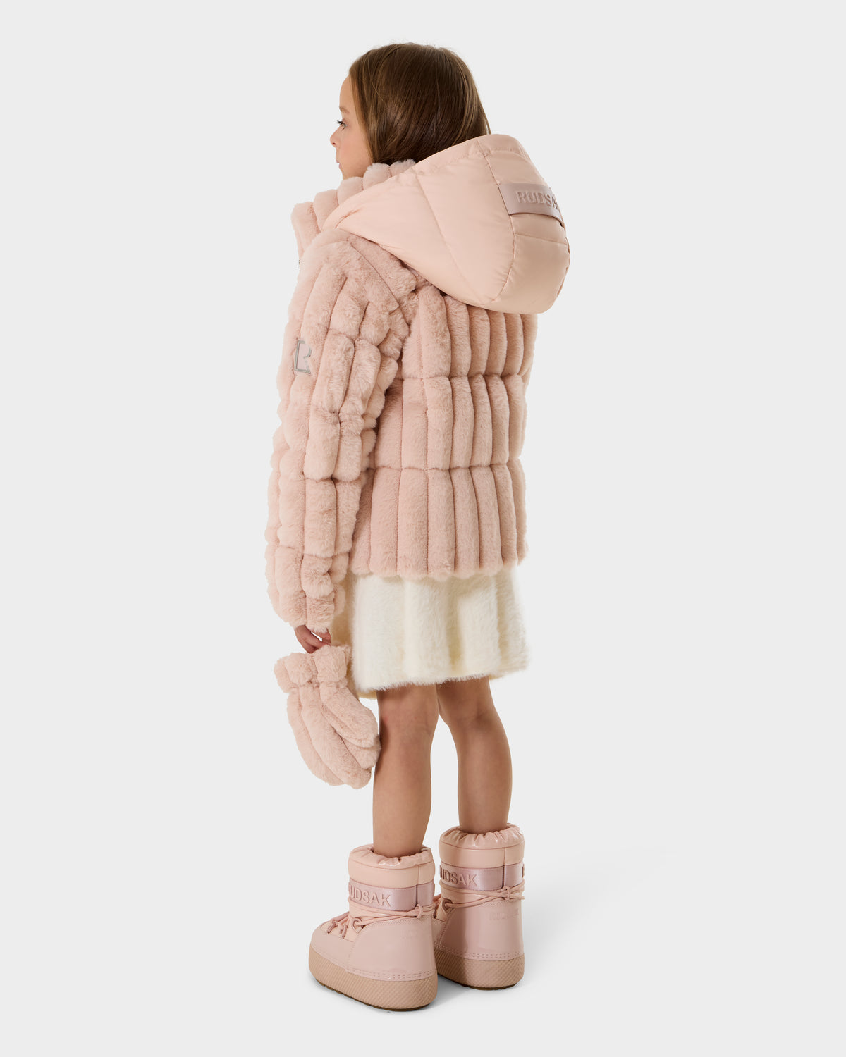 A young girl with long brown hair stands facing away against a white background, wearing the Rudsak ANASTASIA K - 0125000 Pastel Pink puffer jacket, matching mittens, a cream dress, and pink snow boots.