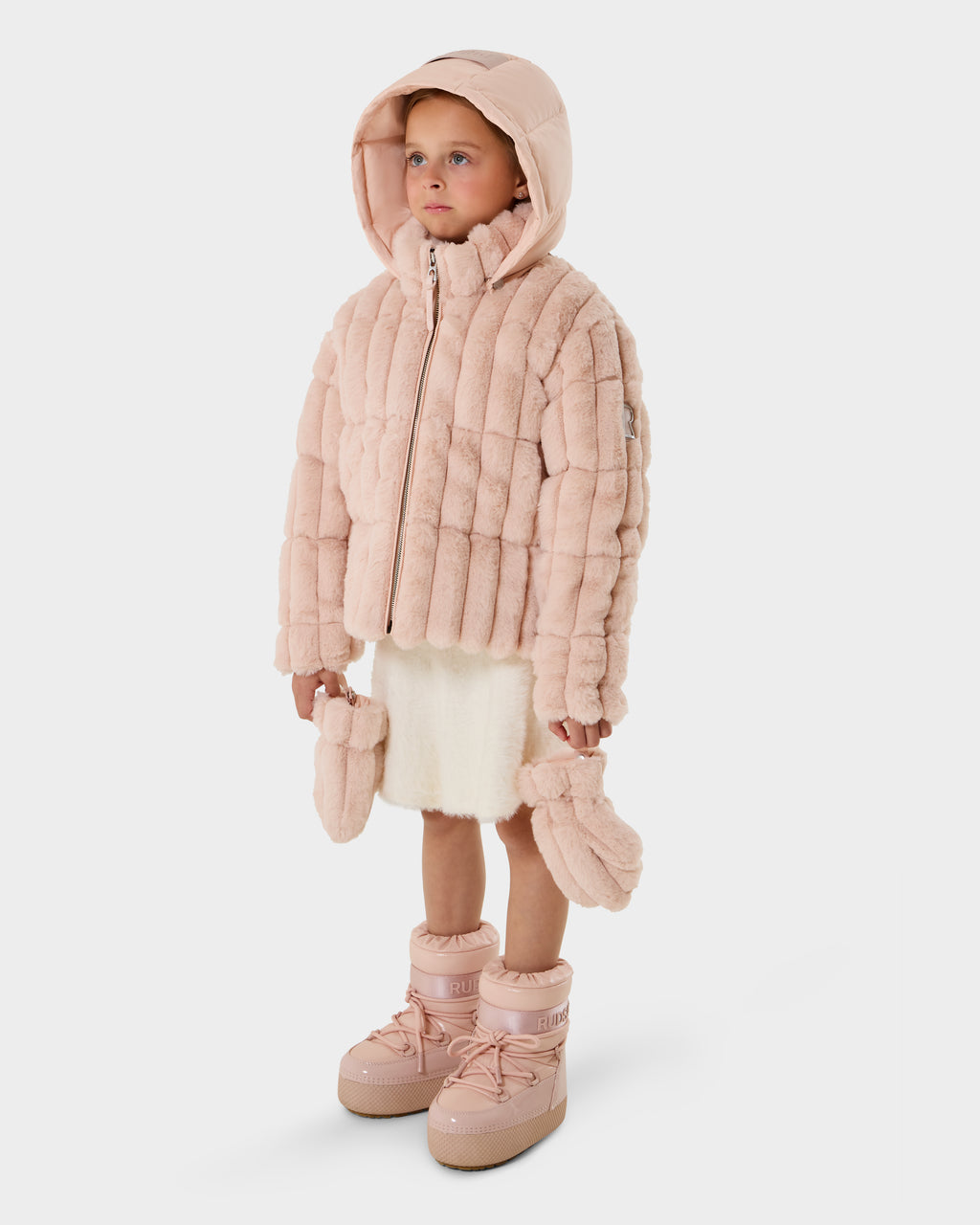 A young child stands against a white background wearing the Rudsak ANASTASIA K - 0125000 Pastel Pink puffer jacket with hood, a cream skirt, pink mittens in hand, and pink winter boots.