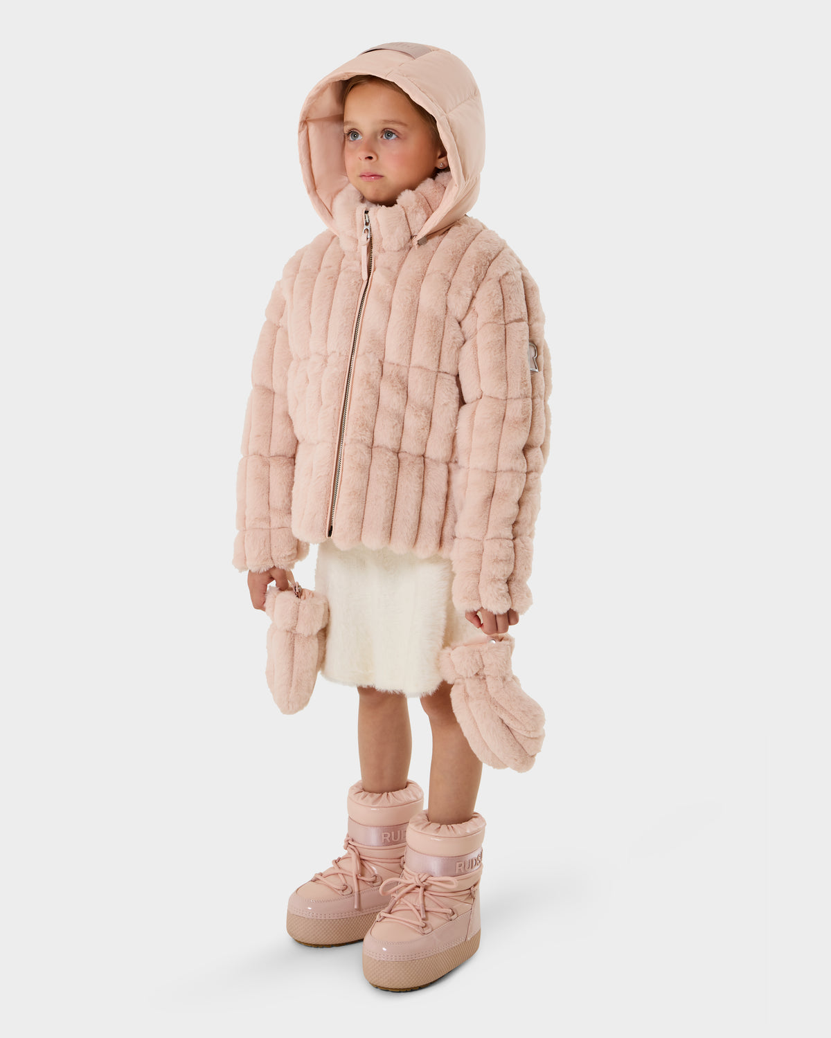 A young child stands against a white background wearing the Rudsak ANASTASIA K - 0125000 Pastel Pink puffer jacket with hood, a cream skirt, pink mittens in hand, and pink winter boots.