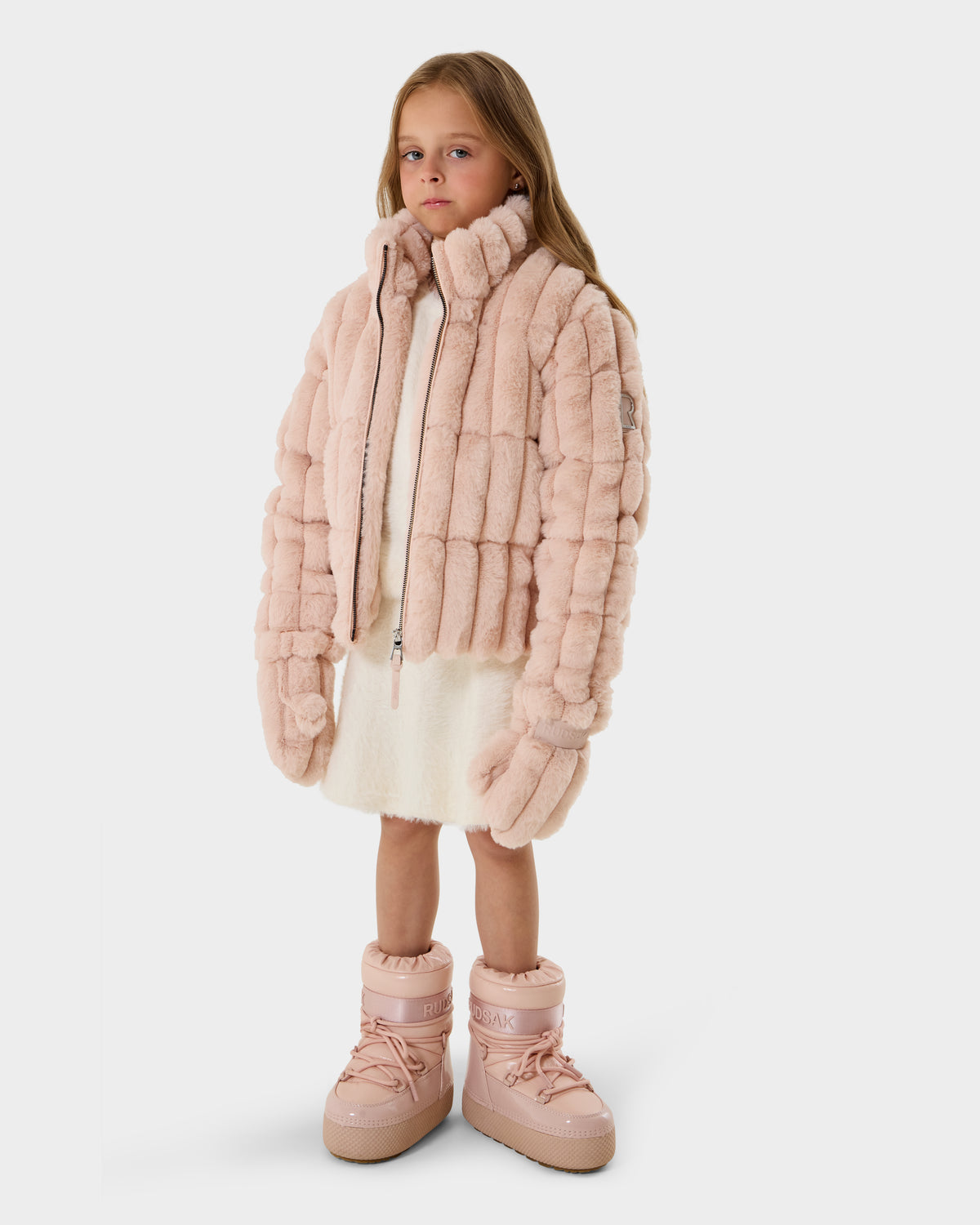 A young girl faces forward in a Rudsak ANASTASIA K - 0125000 Pastel Pink faux fur puffer, white dress, and matching pink boots. She has long straight hair and a neutral expression against a plain white background.