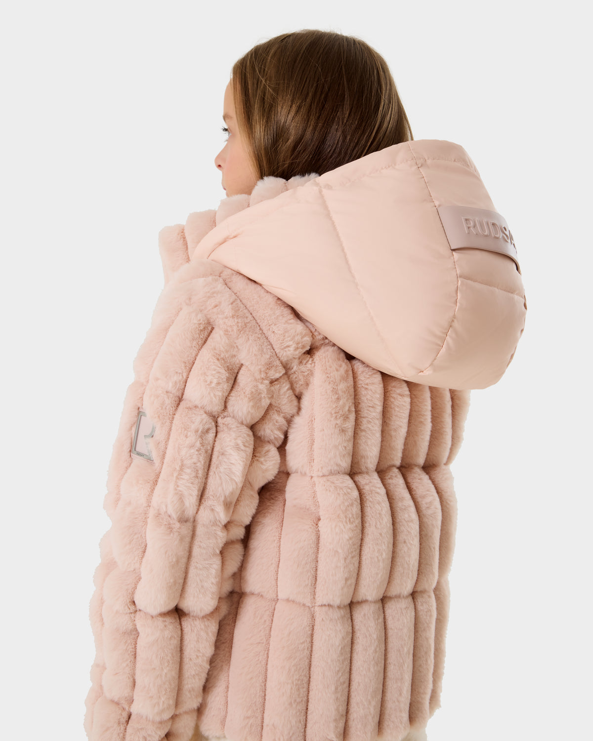 A young person with light brown hair wears the Rudsak ANASTASIA K - 0125000 Pastel Pink plush, hooded jacket. The back view highlights its quilted texture and logo patch on the hood, set against a plain white background.
