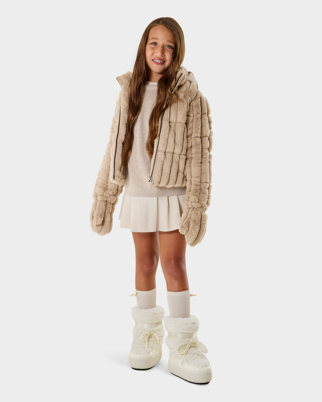 A young girl with long brown hair models the Rudsak ANASTASIA K - 0125000 Vanilla faux fur jacket, paired with a white sweater, cream skirt, knee-high socks, and white boots while smiling against a plain white background.