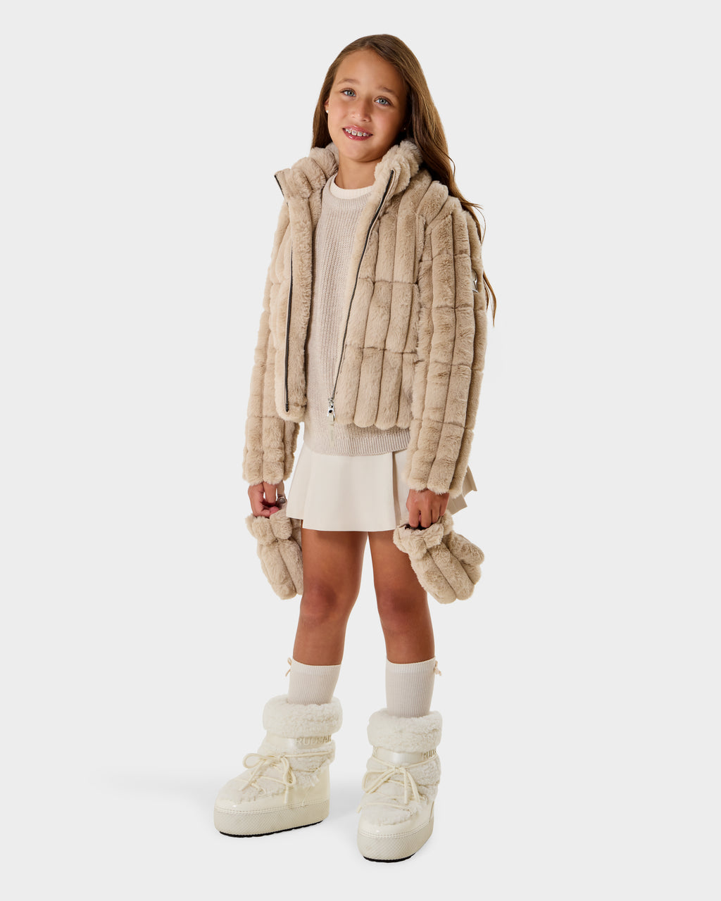 A young girl wearing the Rudsak ANASTASIA K - 0125000 Vanilla jacket smiles against a plain white background, styled with matching mittens, a cream sweater, a white skirt, and winter boots.